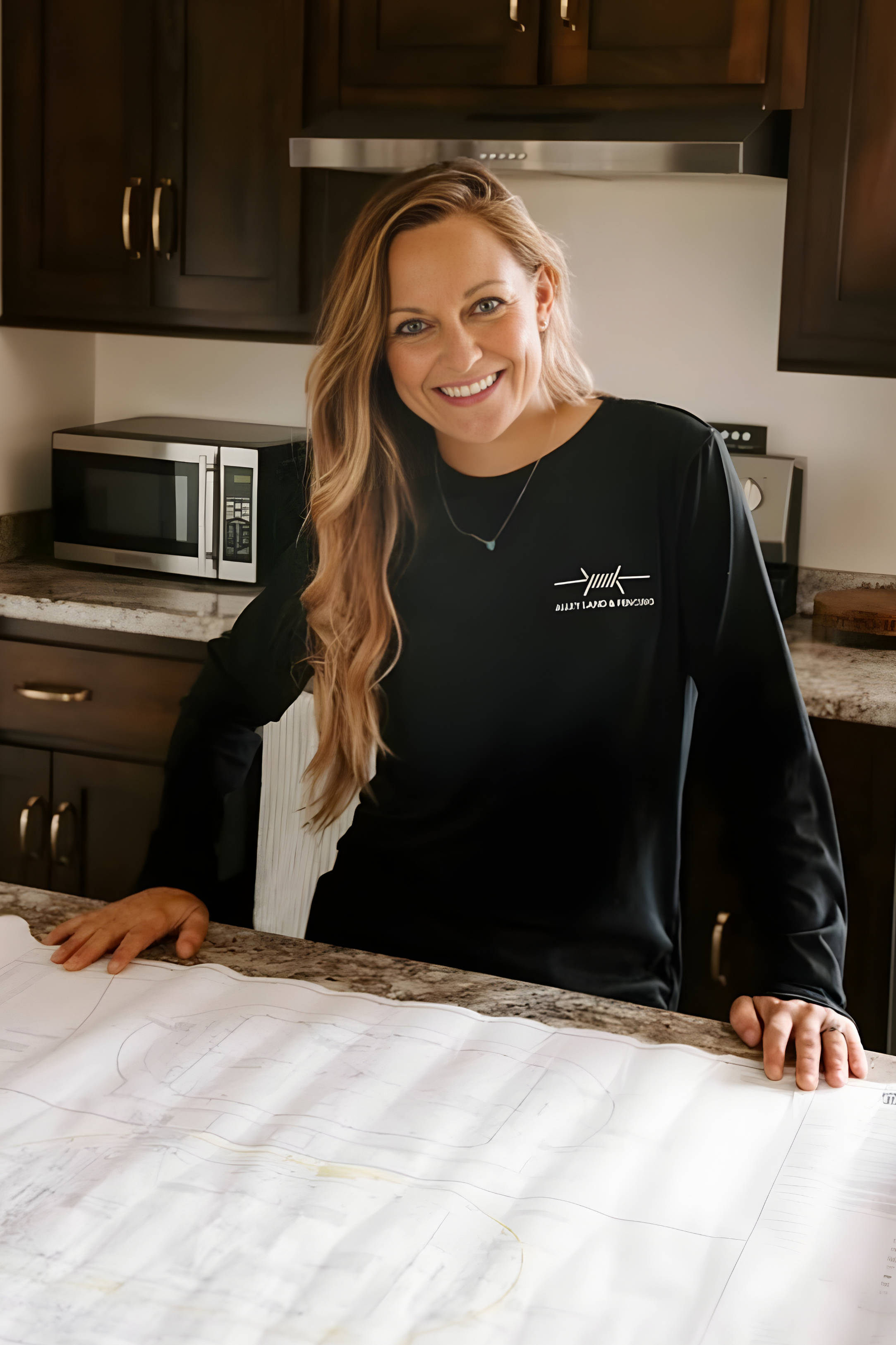 Woman smiling at camera, standing in a kitchen with her hands on a large blueprint or technical drawing laid out on the counter.
