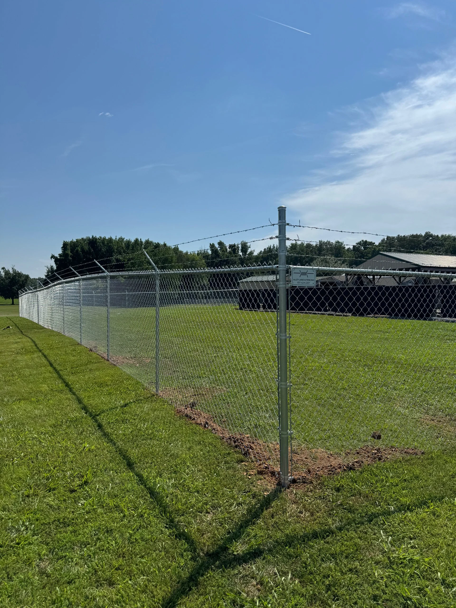 Tall chain-link fence with barbed wire top, set on grass field under blue sky with few clouds.
