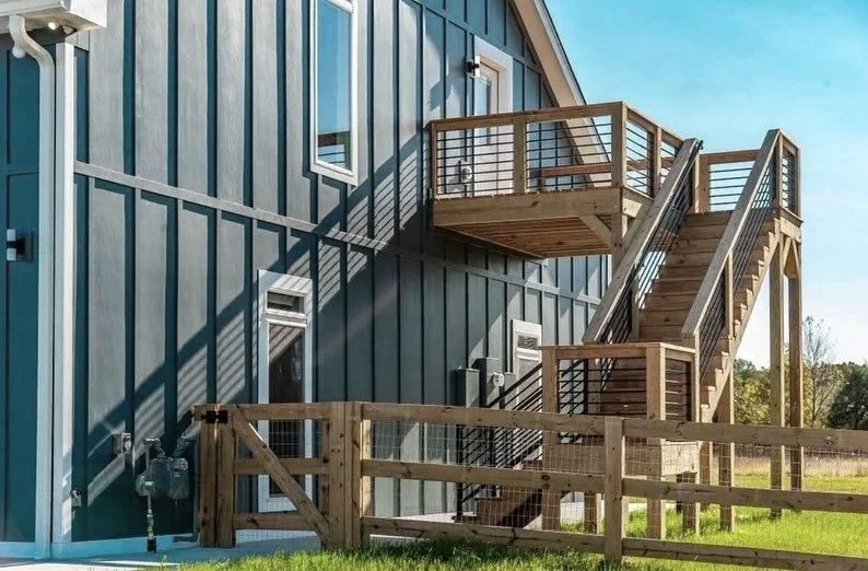 Exterior view of a blue building with multiple outdoor wooden staircases and platforms, sunlight casting shadows, and a grassy field in the background.