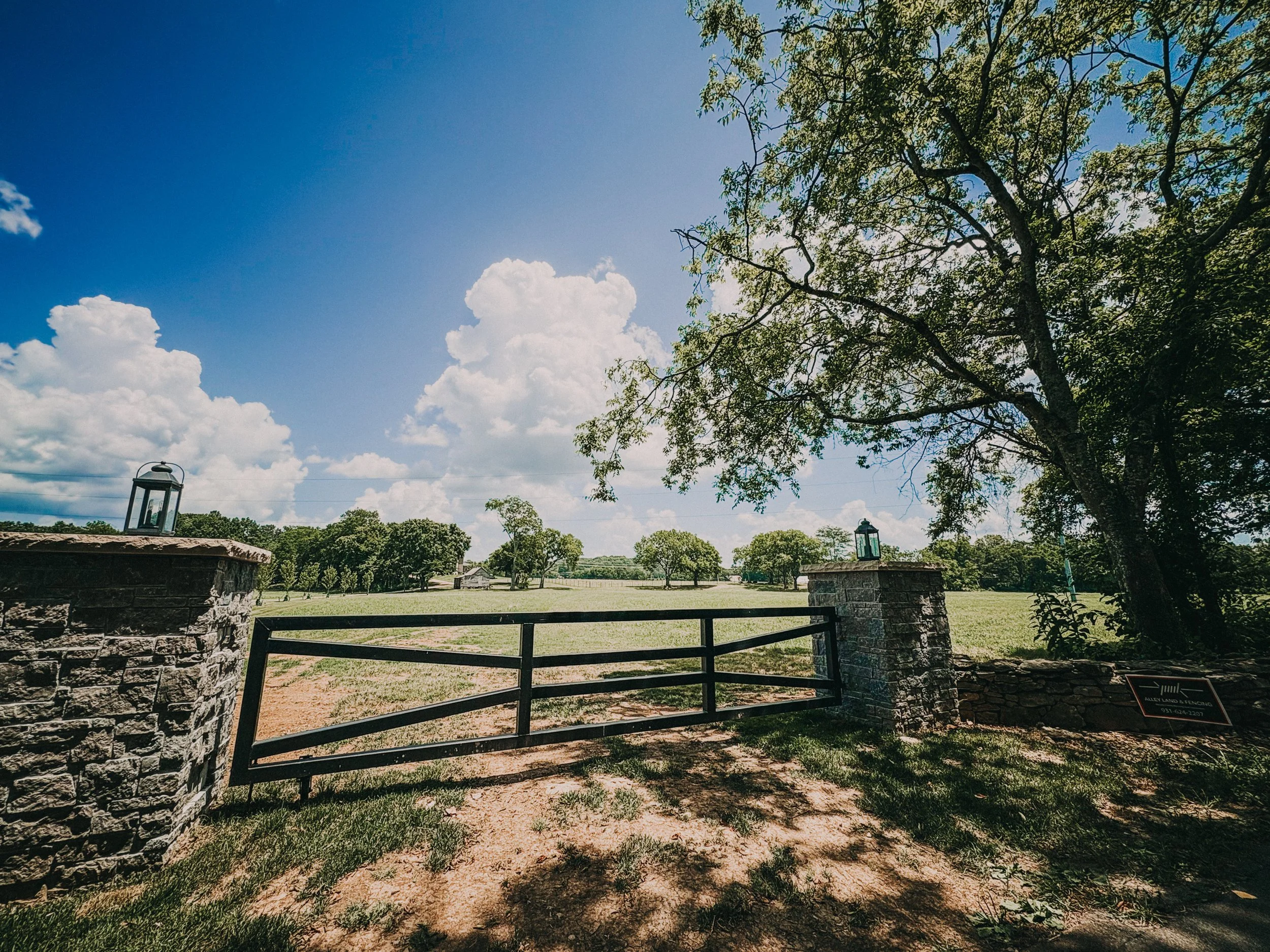 A large open field with trees in the background, a stone and metal gate in the foreground, and a bright blue sky with scattered clouds.