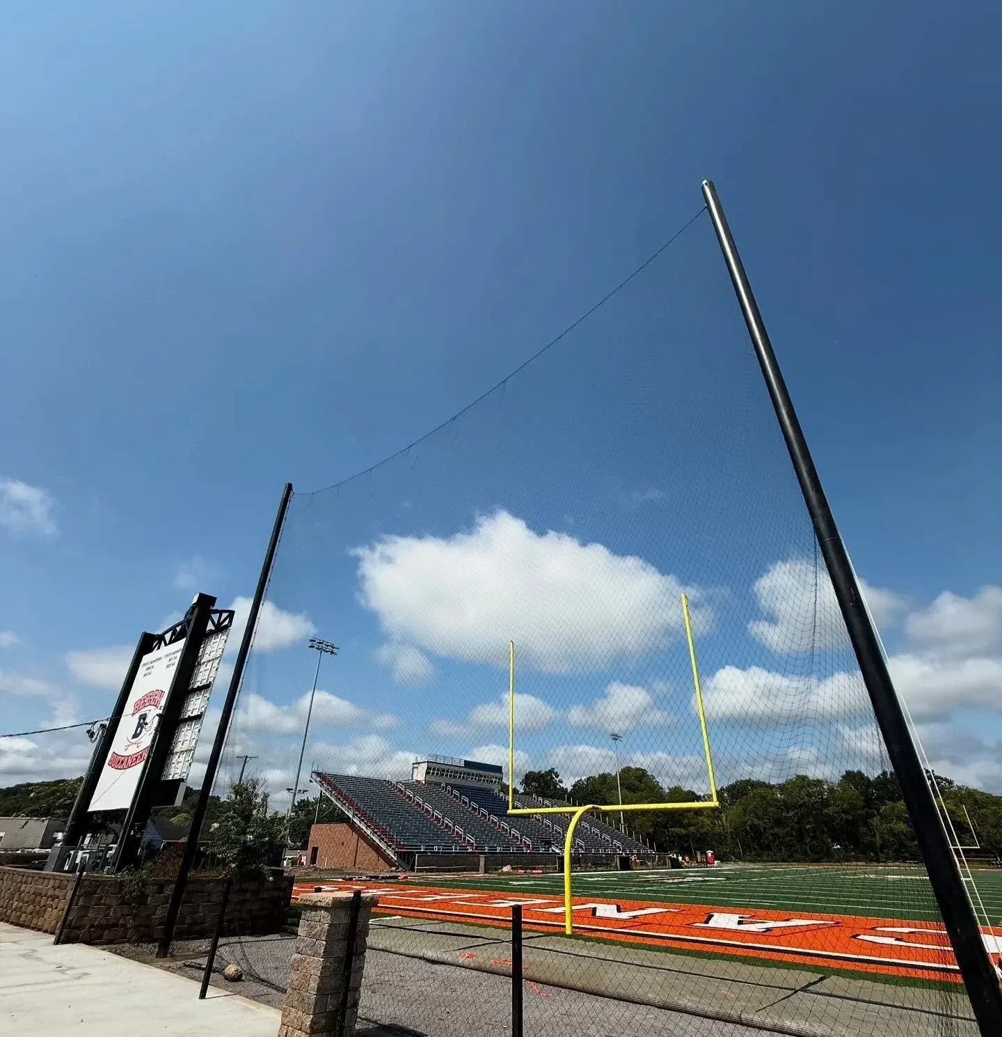 Empty football field with goalposts and stadium bleachers in the background, overcast sky, and a protective netting in the foreground.