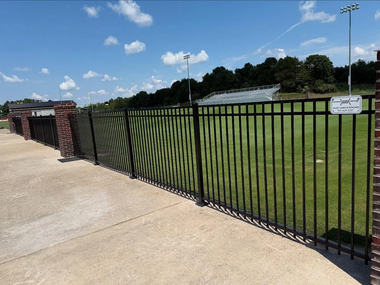Black metal fence with brick pillars along a concrete sidewalk, enclosing a grassy sports field with bleachers under a blue sky with scattered clouds.