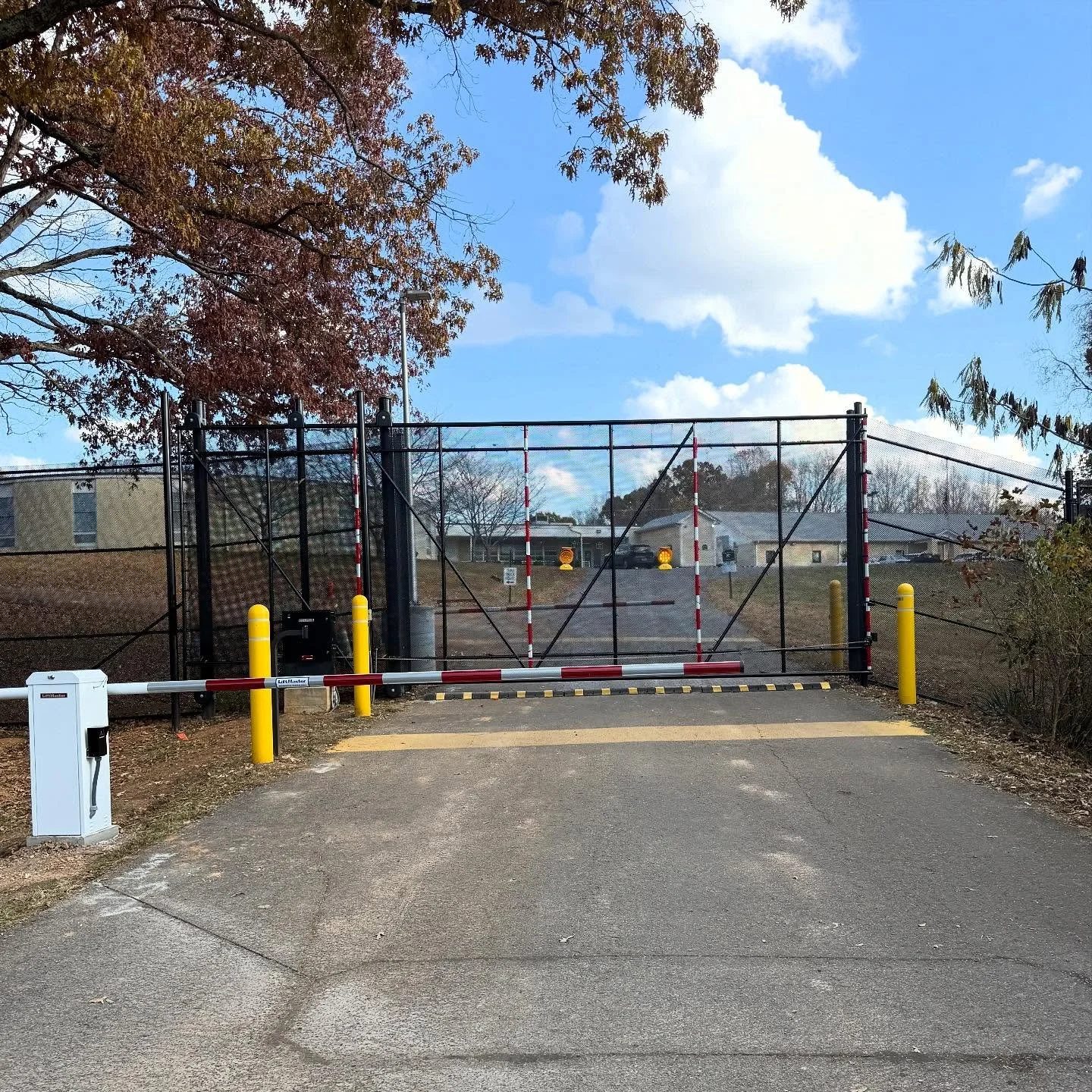 A fenced gate blocking vehicle access to a parking lot, with a barrier arm, yellow bollards, and an access control panel on the left. There are trees with autumn leaves, a blue sky with some clouds, and buildings in the background.