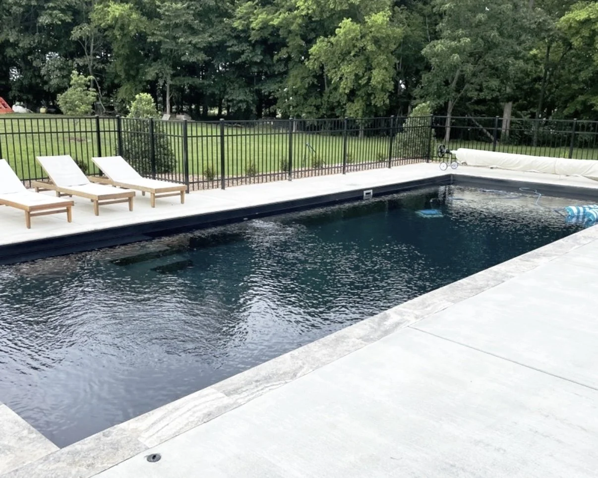 A rectangular in-ground swimming pool with dark water, surrounded by a light-colored stone deck, with three white lounge chairs along the fence, a grassy yard and trees in the background.