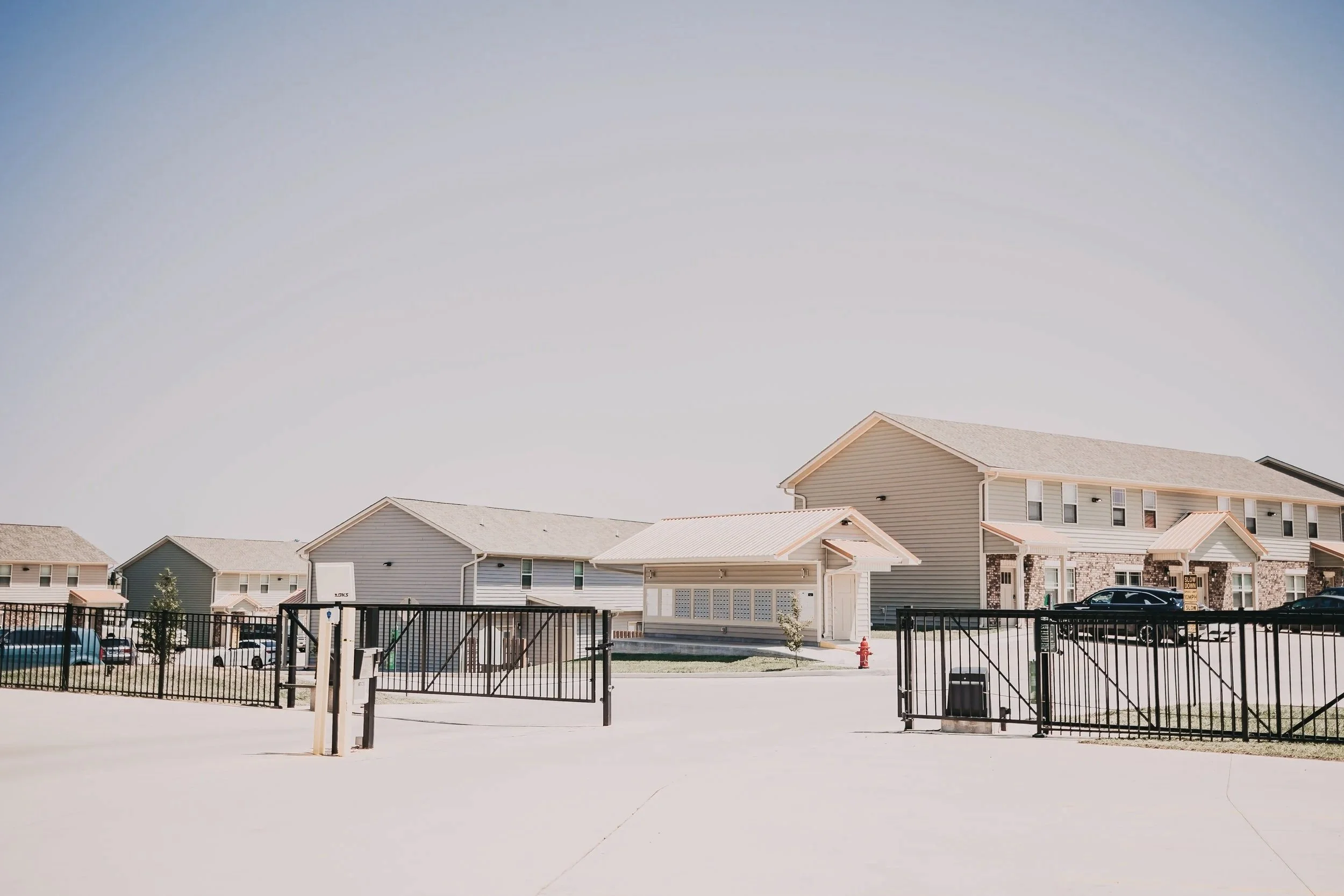 A gated residential community with houses, parked cars, and a small building in the center under a clear sky.