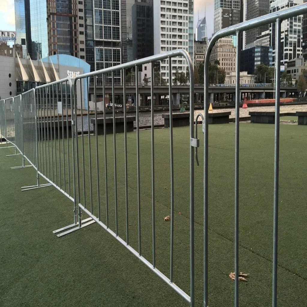 Metal crowd control barriers-guarding an outdoor artificial turf area in a city with tall buildings and a river in the background.