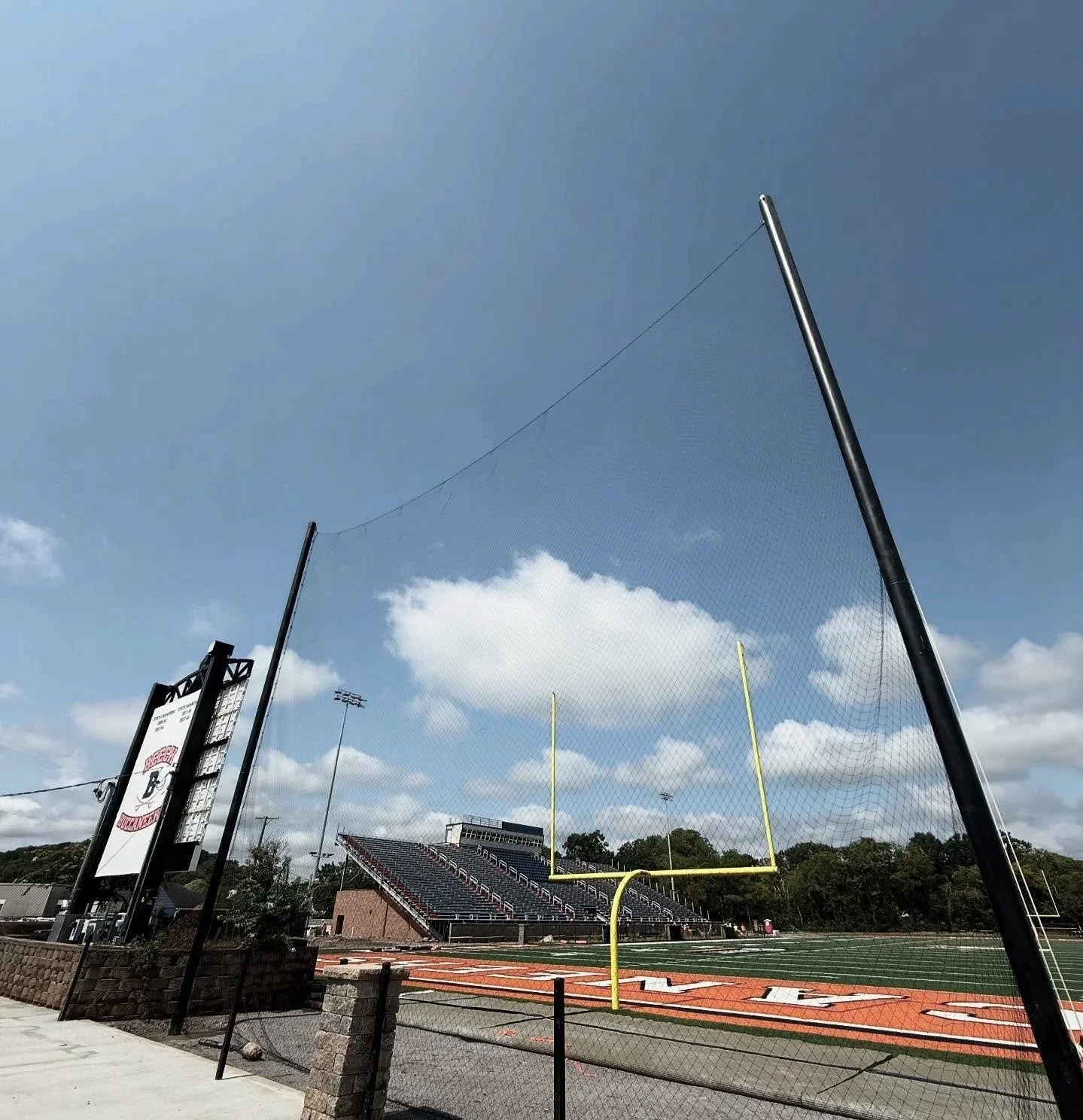 Empty football field with goalposts and stadium bleachers in the background, overcast sky, and a protective netting in the foreground.
