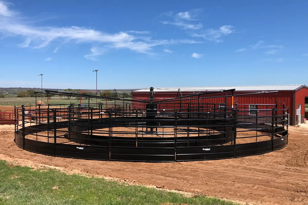 A round horse riding arena with black fencing, located outdoors under a blue sky with clouds, with a red barn in the background.