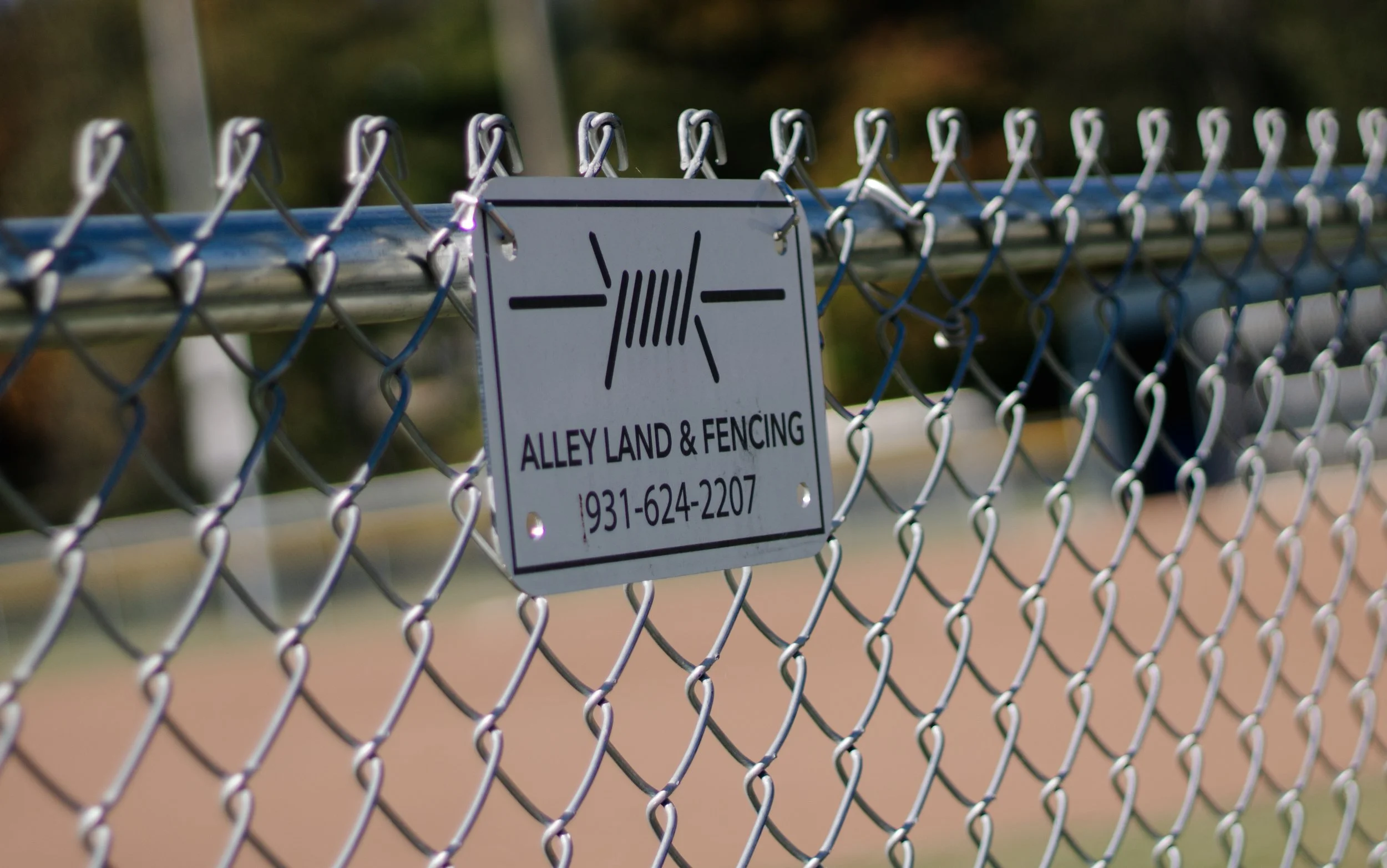 Close-up of a chain-link fence with a sign that reads "ALLEY LAND & FENCING" and a phone number, attached to the fence.