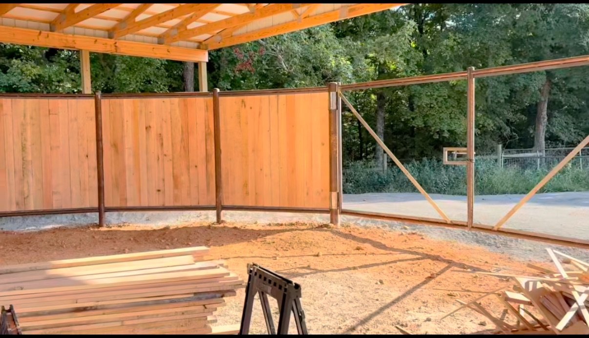 Construction site with a partially built wooden fence and gate, surrounded by trees and a dirt ground.