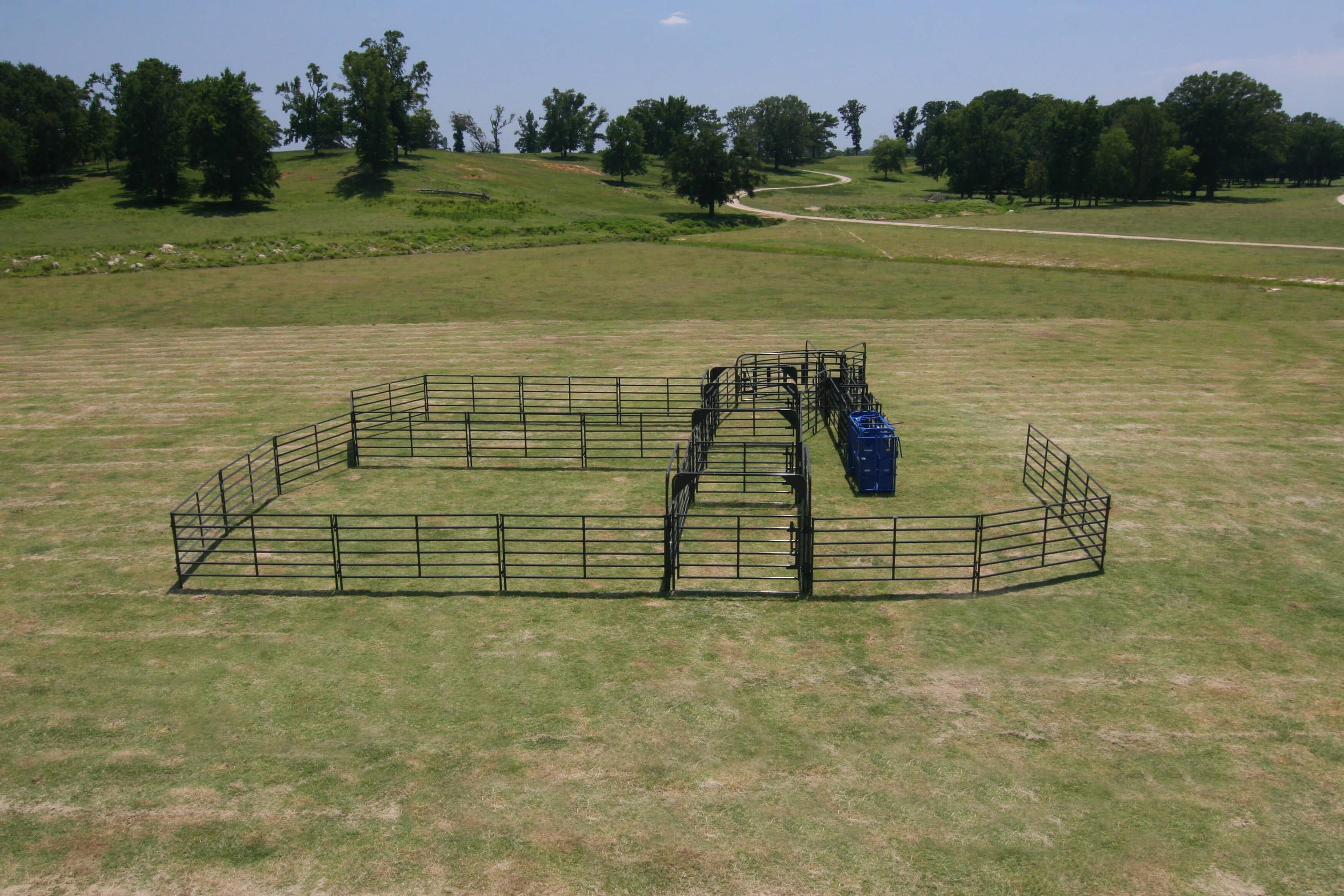 An empty outdoor animal pen on a grassy field with a blue livestock manager and black metal fencing, surrounded by green rolling hills and a dirt road in the distance.