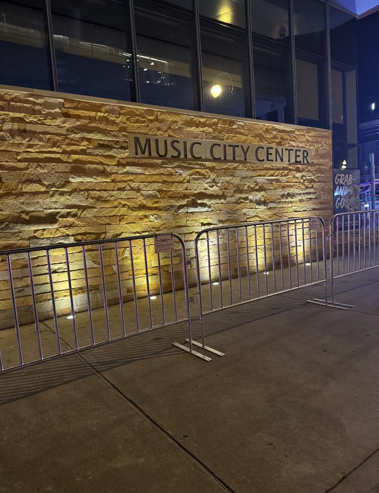 Nighttime exterior view of the Music City Center, a building with stone and glass walls, illuminated by yellow lights, with signs reading 'Music City Center' and 'Grab and Go', and temporary fencing barricades in the foreground.