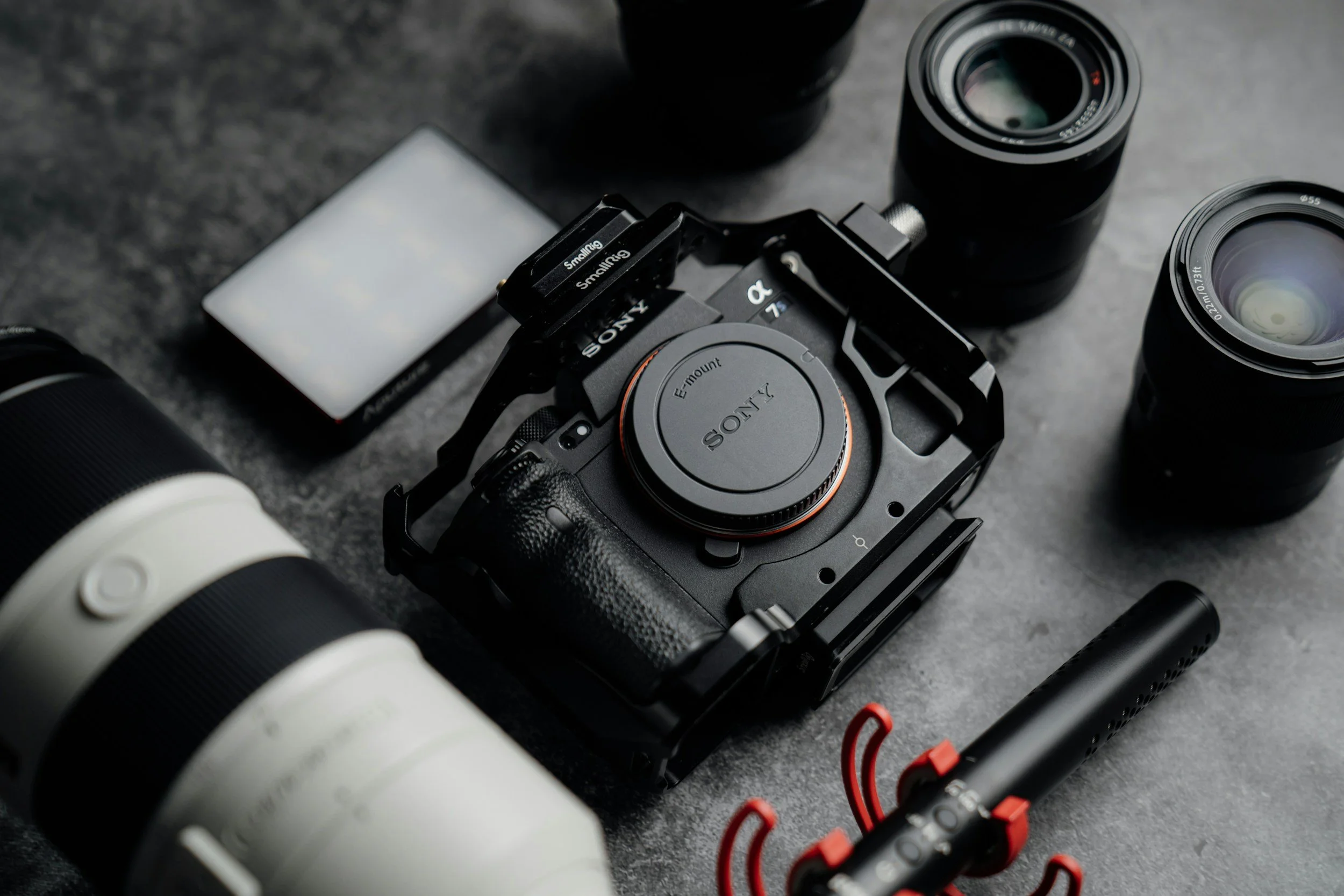 Camera equipment including a Sony mirrorless camera with lens attached, two additional camera lenses, a camera flash, a smartphone, and a red and black LED light on a grey surface.