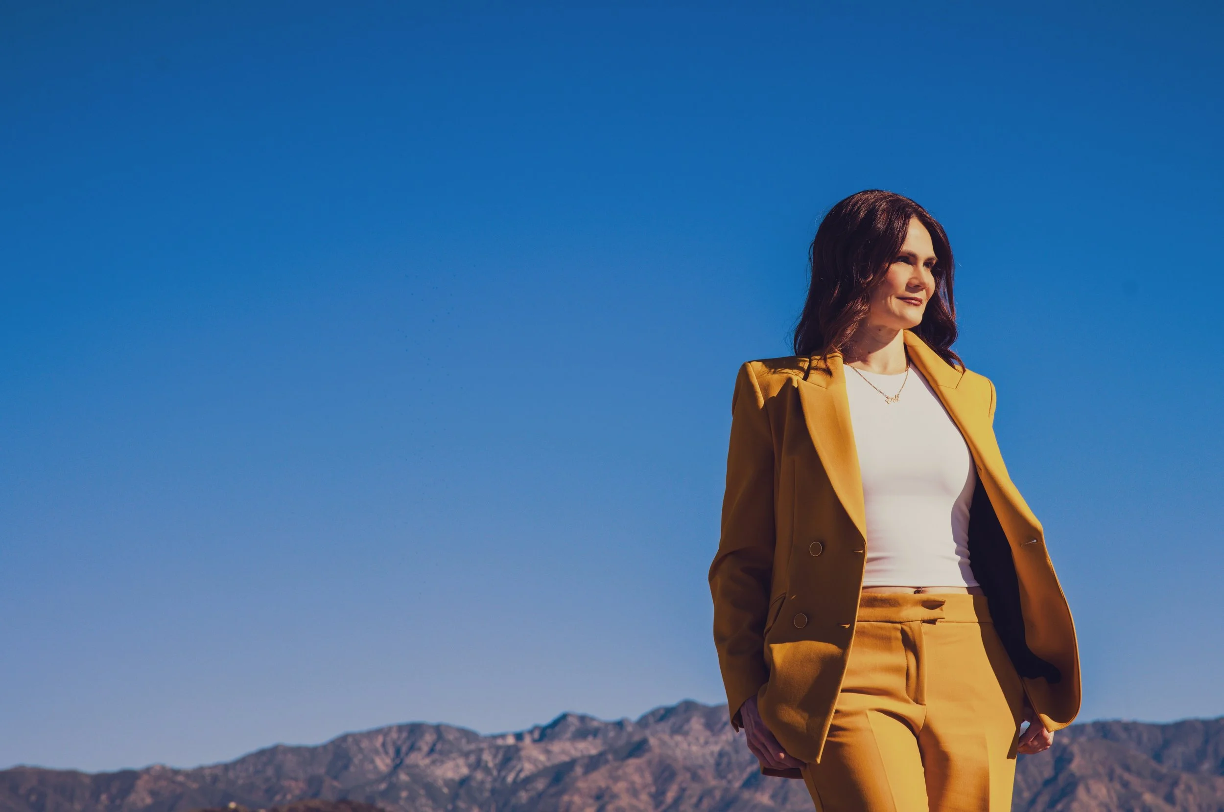 A woman in a yellow suit standing outdoors with mountains in the background under a clear blue sky.
