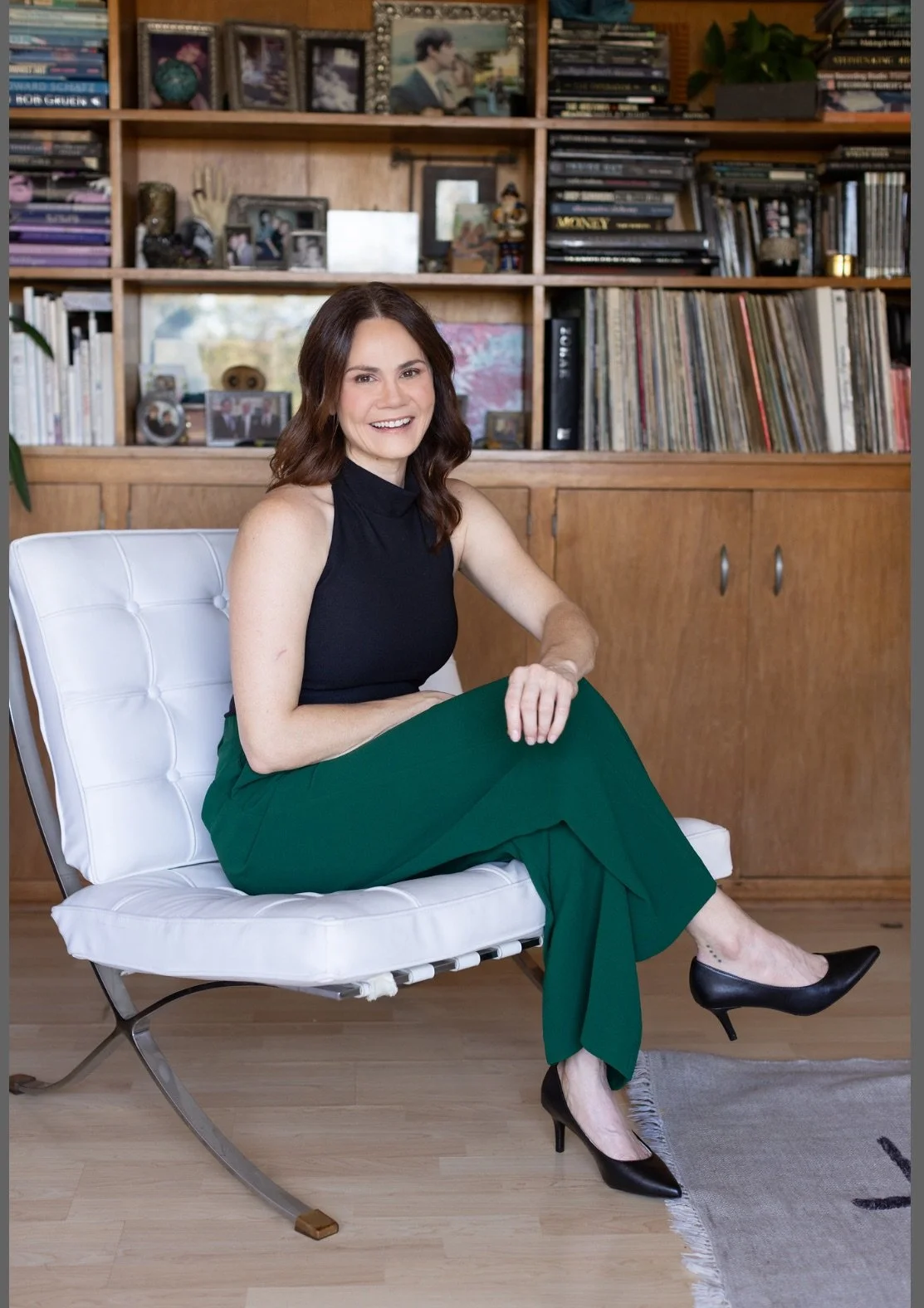 Anxiety, OCD therapist in Los Angeles with shoulder-length brown hair, smiling, sitting on a modern white chair with a wooden and bookshelf background. She is wearing a black sleeveless top, green pants, and black high heels.