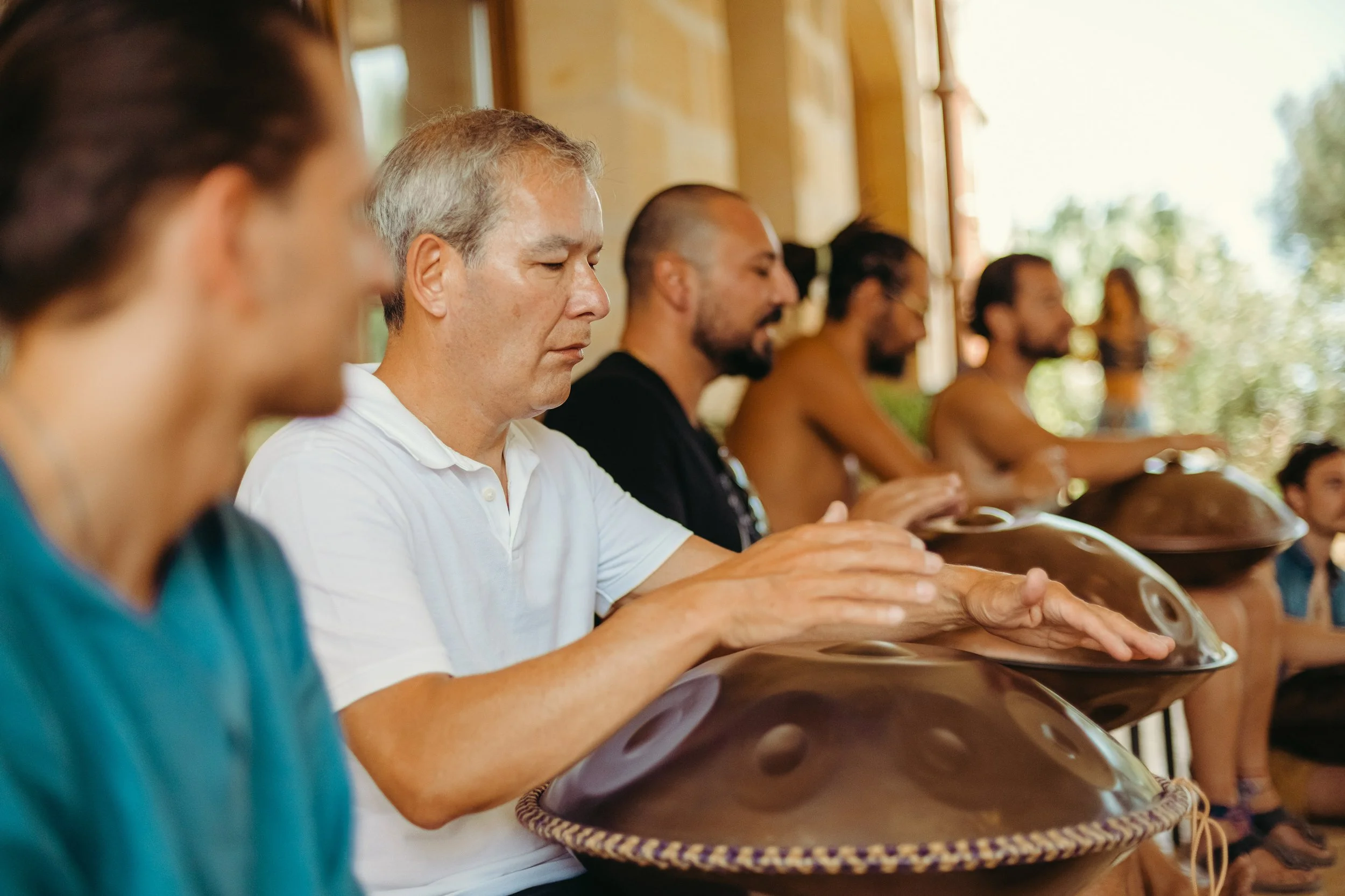 A group of people sitting on a wooden bench playing handpan drums during a music session indoors with natural light.
