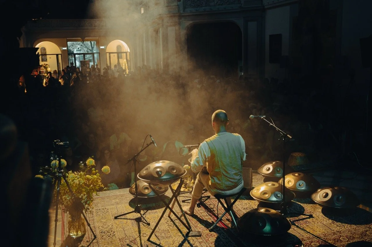 A musician playing the hang drum on stage with a microphone and multiple hang drums around him, a flower arrangement, and an audience in the background.