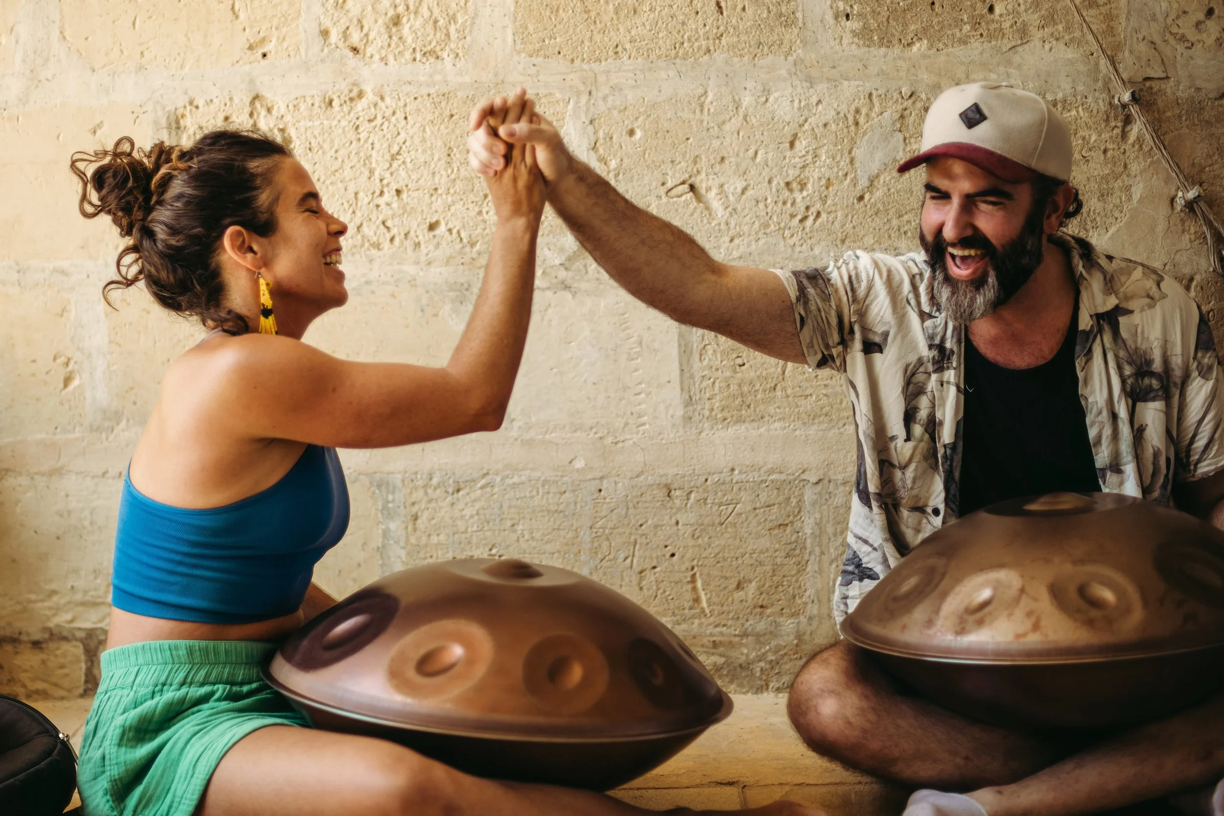 A woman and a man are sitting on the floor playing hang drums. They are smiling at each other and high-fiving. The woman is wearing a blue crop top and green shorts. The man is wearing a patterned shirt, baseball cap, and has a beard. They are sitting in front of a textured stone wall.