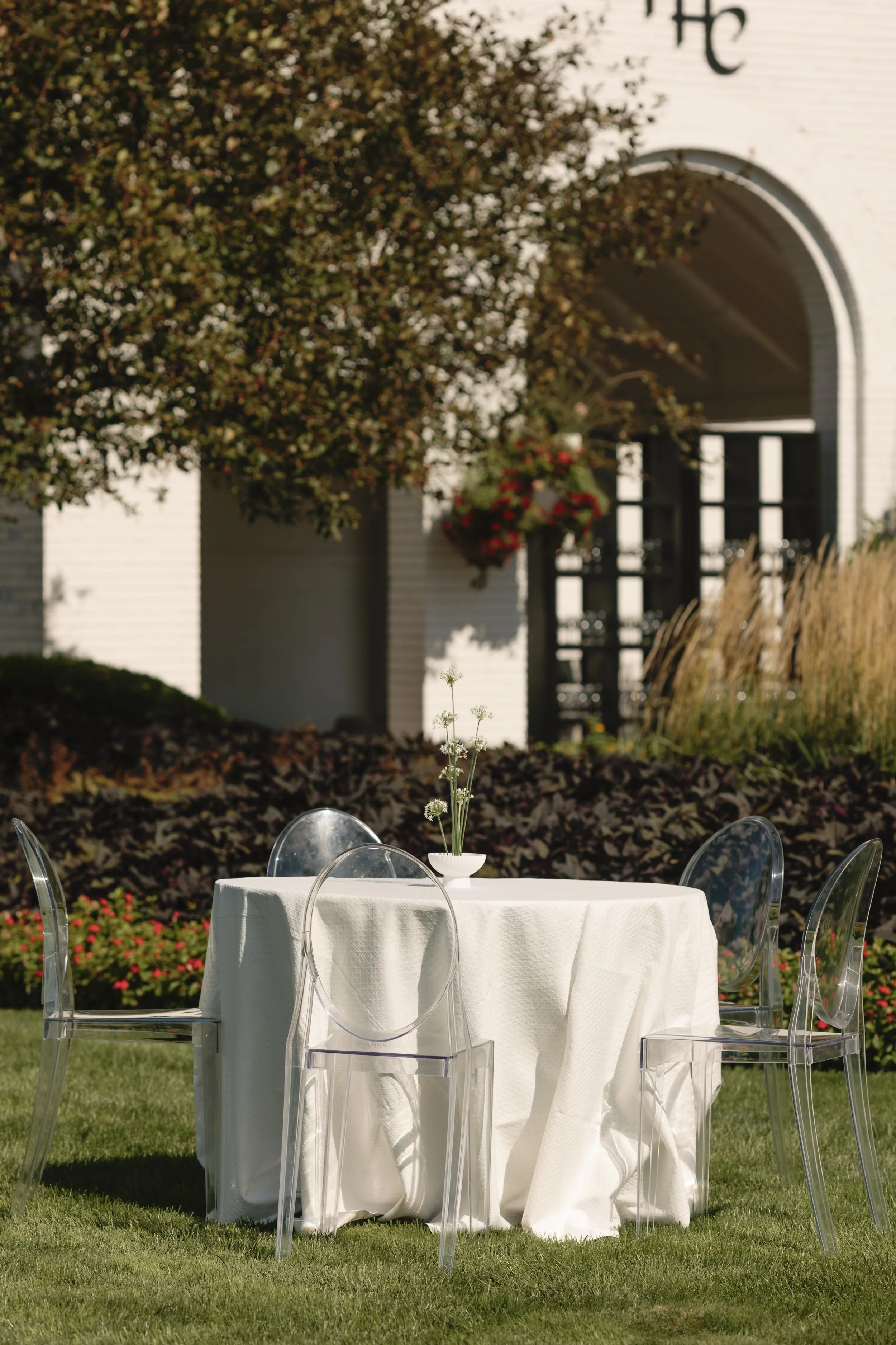 Outside garden scene with a round table draped in a white cloth, set with a small vase of white flowers, surrounded by clear modern chairs. In the background, there are trees, plants, and part of a white building with an arched entrance and hanging f