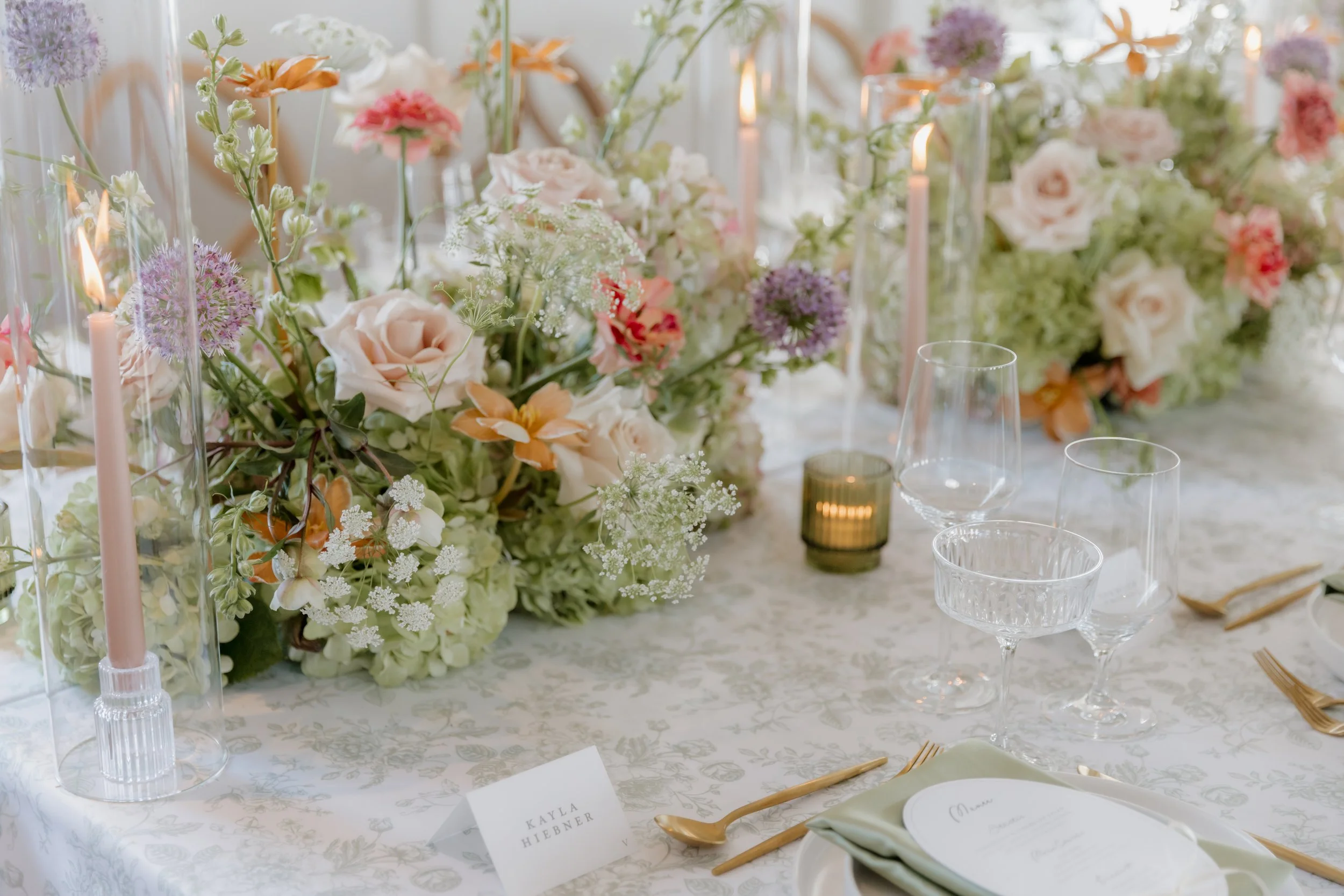A wedding reception table decorated with a floral centerpiece, candles, glassware, and place settings.