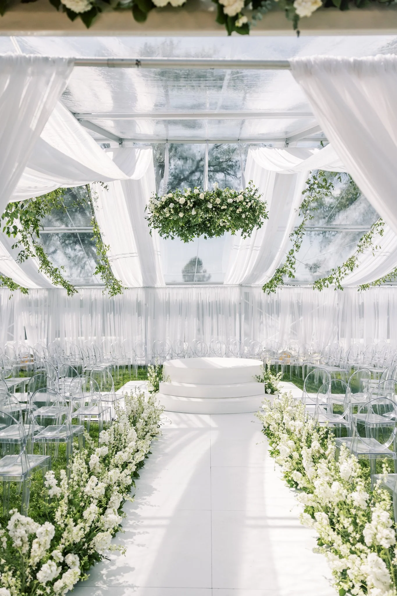 Elegant white wedding altar inside a glass tent with draped white fabric, floral arrangements, and clear acrylic chairs for guests.