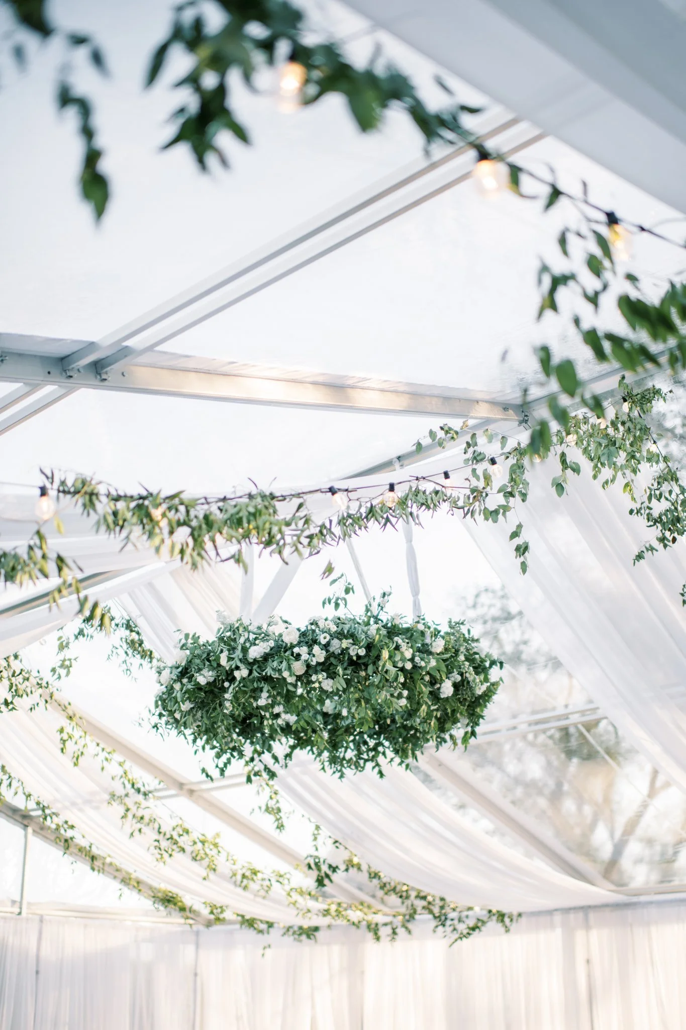 Hanging floral arrangement with green leaves and white flowers inside a tent decorated with string lights and draped white fabric.