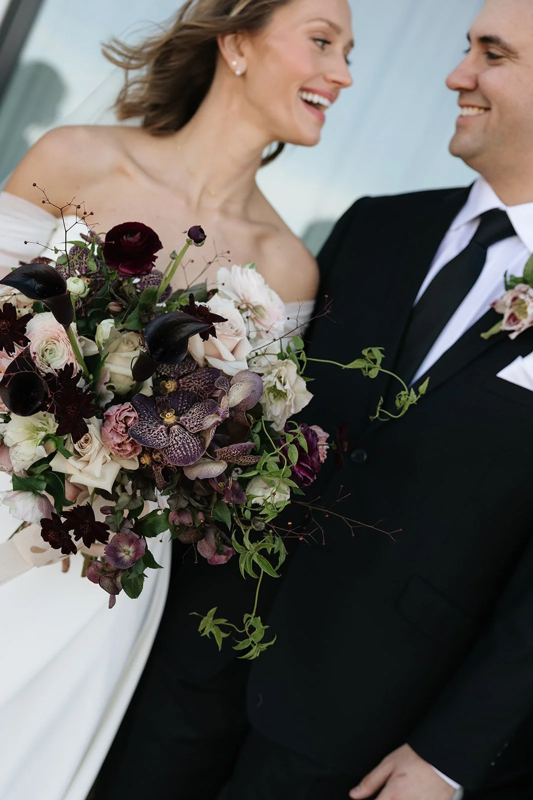 A bride and groom smiling at each other, with the bride holding a large bouquet of mixed flowers including roses and orchids.