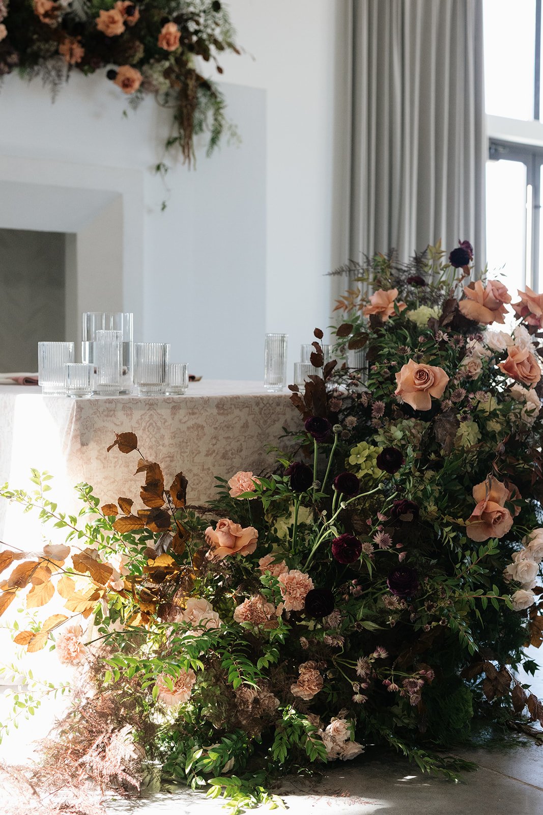 A large floral arrangement with pink roses, dark purple flowers, and green foliage in front of a table with clear glass stemware, inside a room with white walls, curtains, and a fireplace with a floral decoration.