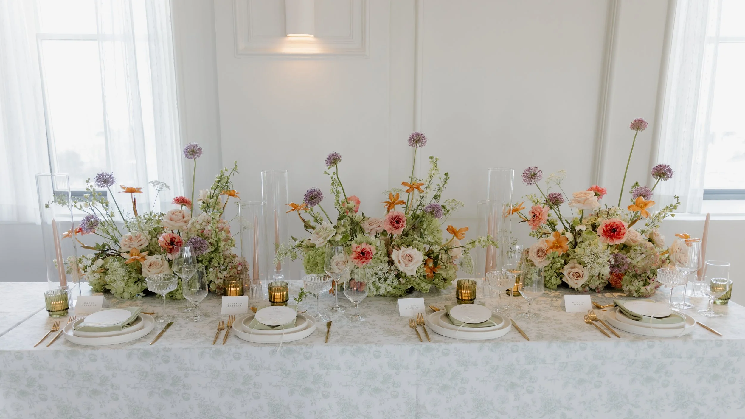 Elegant table setting with three floral centerpieces featuring roses, hydrangeas, and other flowers, glassware, candles, and place cards in a bright, white room with large windows.