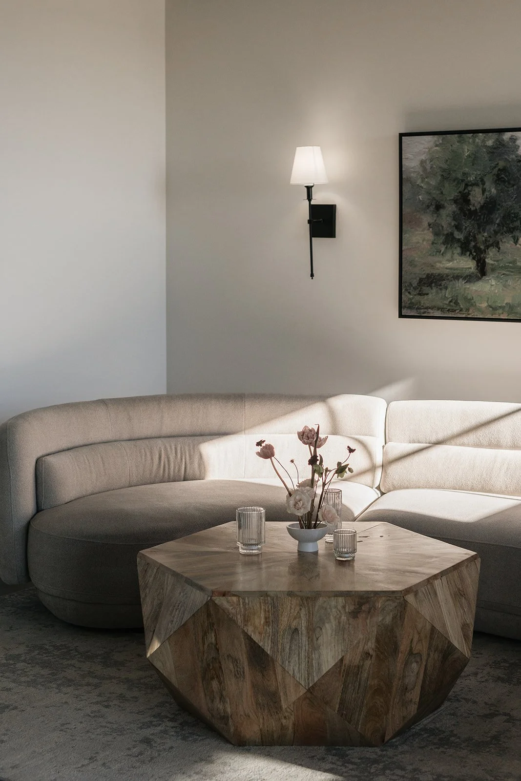 Living room with a beige sectional sofa, a wooden geometric coffee table with vases, a wall-mounted lamp, and a painting on a white wall.