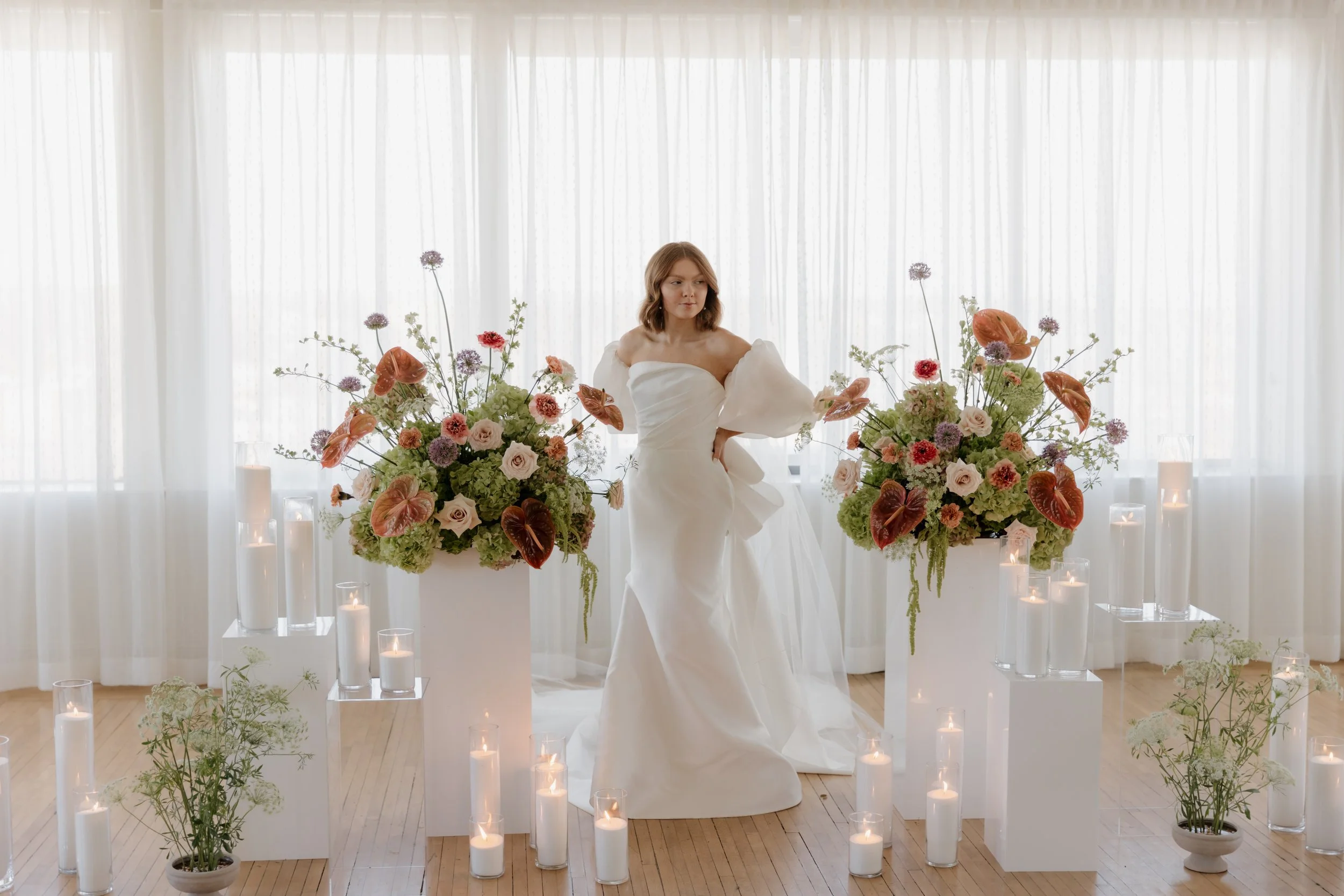 A woman in a white wedding dress standing amidst large floral arrangements and candles in a bright, airy room with sheer white curtains.