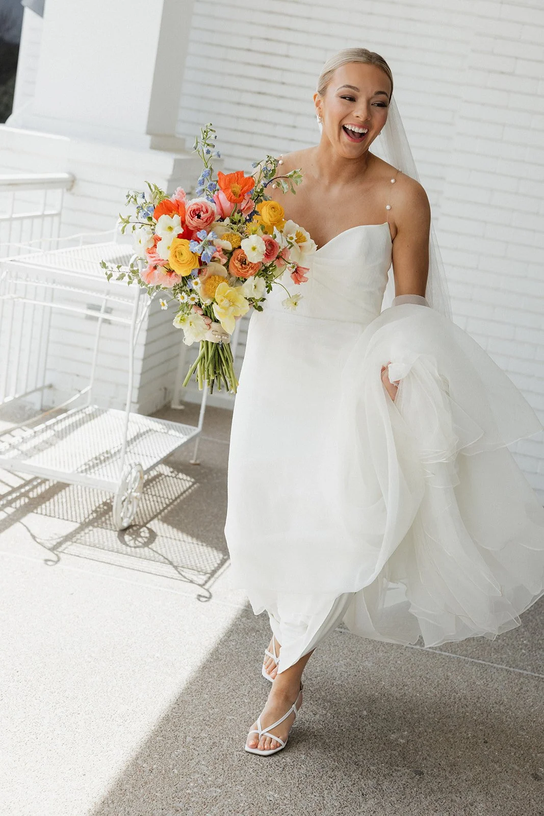 A bride in a white wedding dress holding a colorful bouquet of flowers, smiling and walking outdoors.