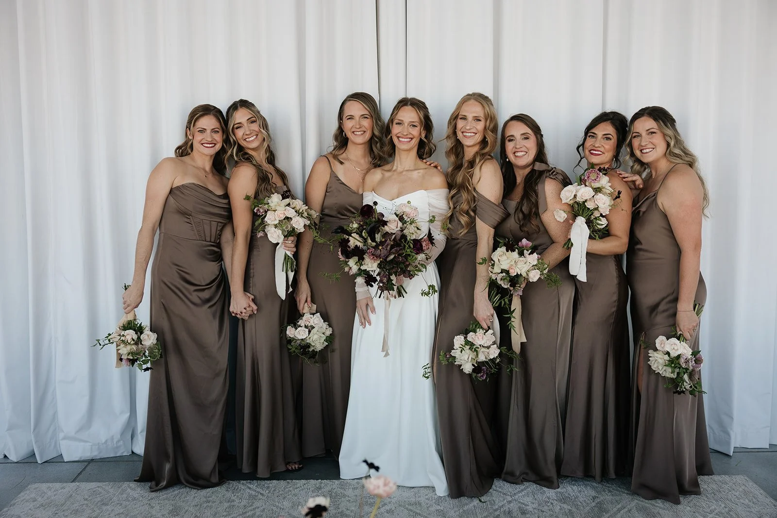 Bridal party of eight women in matching taupe dresses, with the bride in a white gown, holding bouquets of white and dark purple flowers, standing in front of a white curtain.