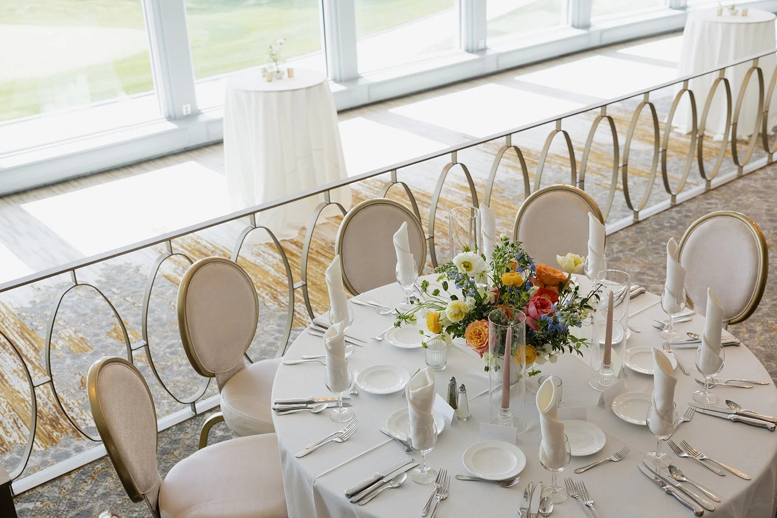 A round banquet table set for a formal event with white tablecloth, neatly folded white napkins, plates, silverware, glasses, and a colorful flower centerpiece. The setup is near large windows allowing natural light.