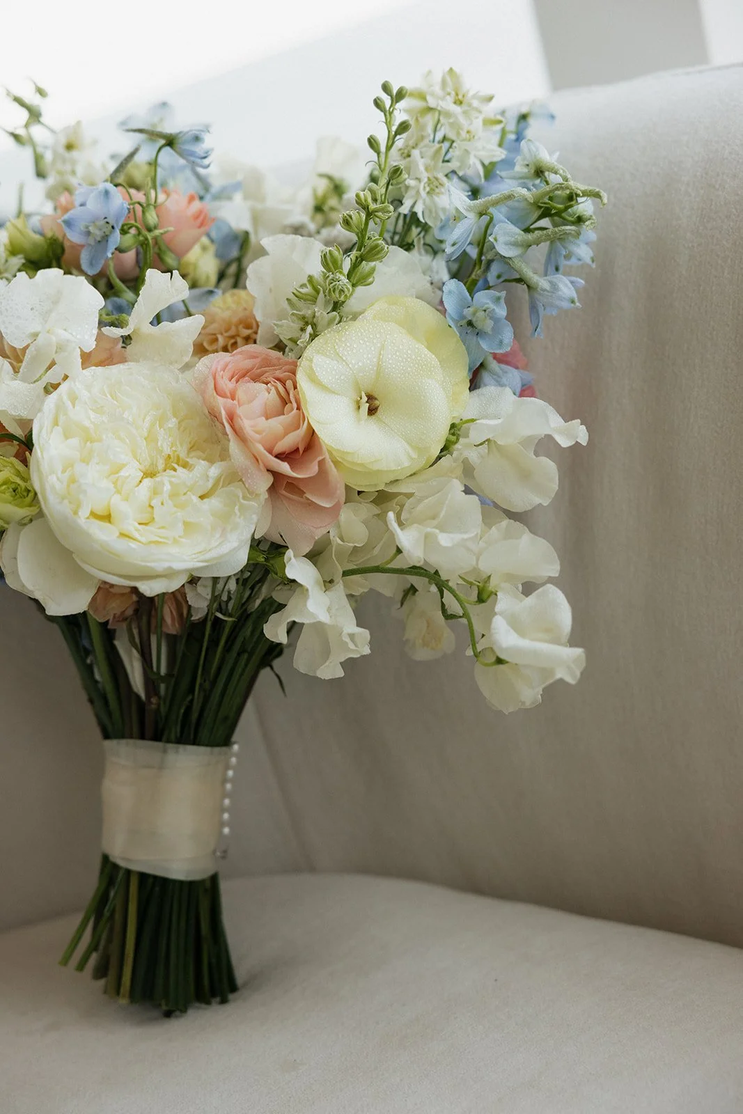 A bouquet of pastel-colored flowers including roses, peonies, and delphiniums, wrapped with a satin ribbon, resting on a light-colored surface with a neutral background.