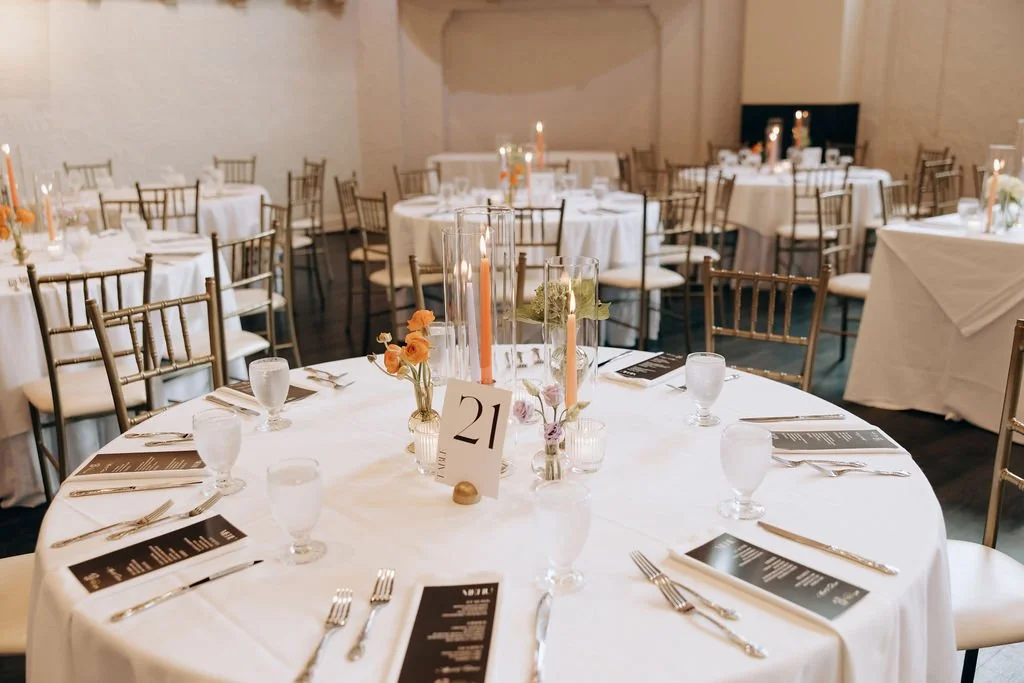 Round banquet table set for a formal event with white tablecloth, floral centerpieces, and place settings including silverware, water glasses, and printed menus. Multiple similar tables and gold chairs are visible in the background.