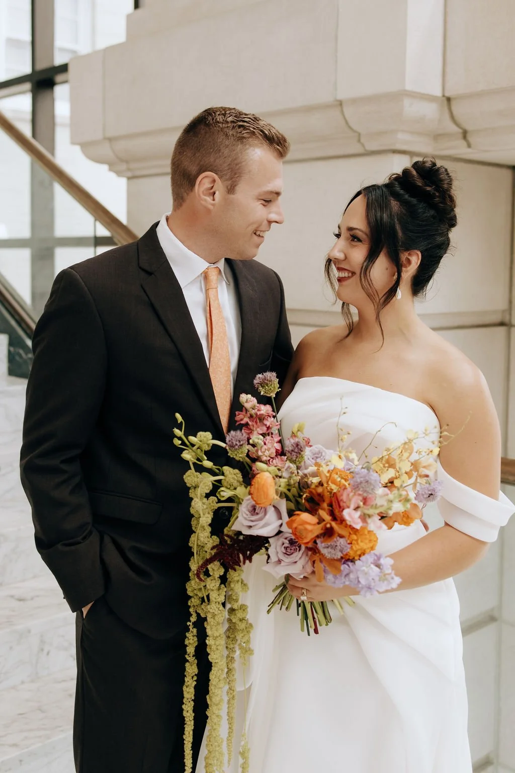 A bride and groom on their wedding day, smiling at each other indoors with a marble staircase in the background. The bride holds a colorful bouquet of flowers.