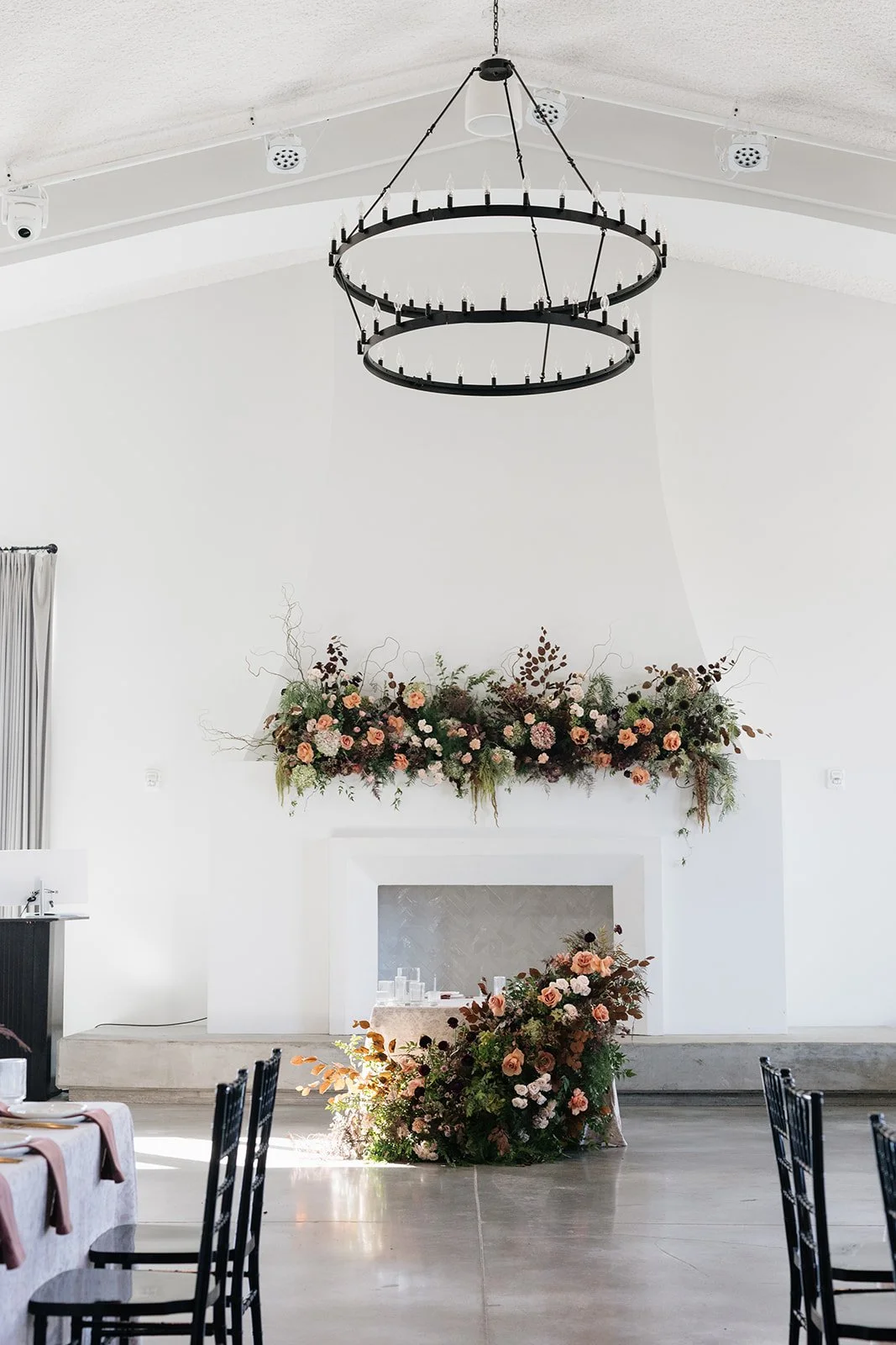 Elegant indoor event space with white walls, high ceiling, and modern black circular chandelier. A decorative floral arrangement is placed above a white fireplace, and another bouquet sits on the floor nearby. Tables with black chairs are set up for 