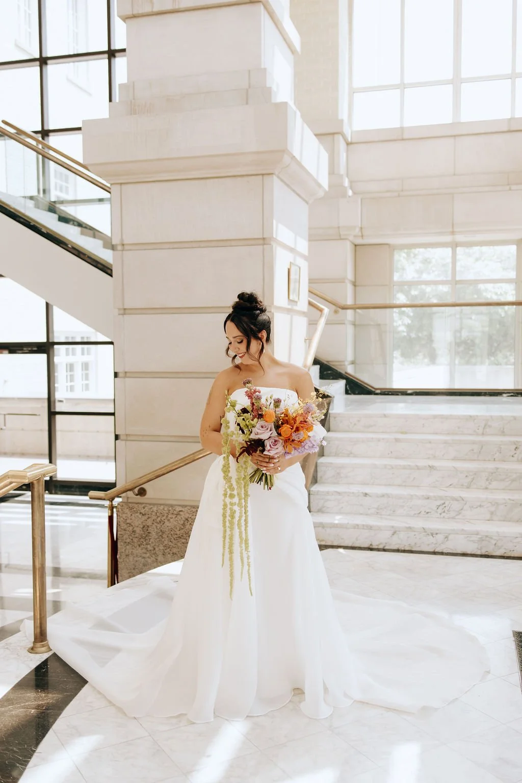 A bride in a strapless white wedding gown holding a bouquet of colorful flowers, standing indoors near a marble staircase with large windows in the background.