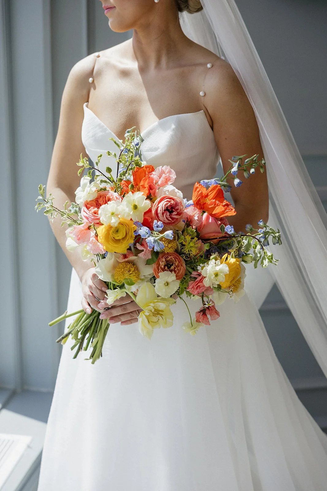 A woman in a white wedding dress holding a colorful bouquet of flowers, with a veil on her head, standing by a window.