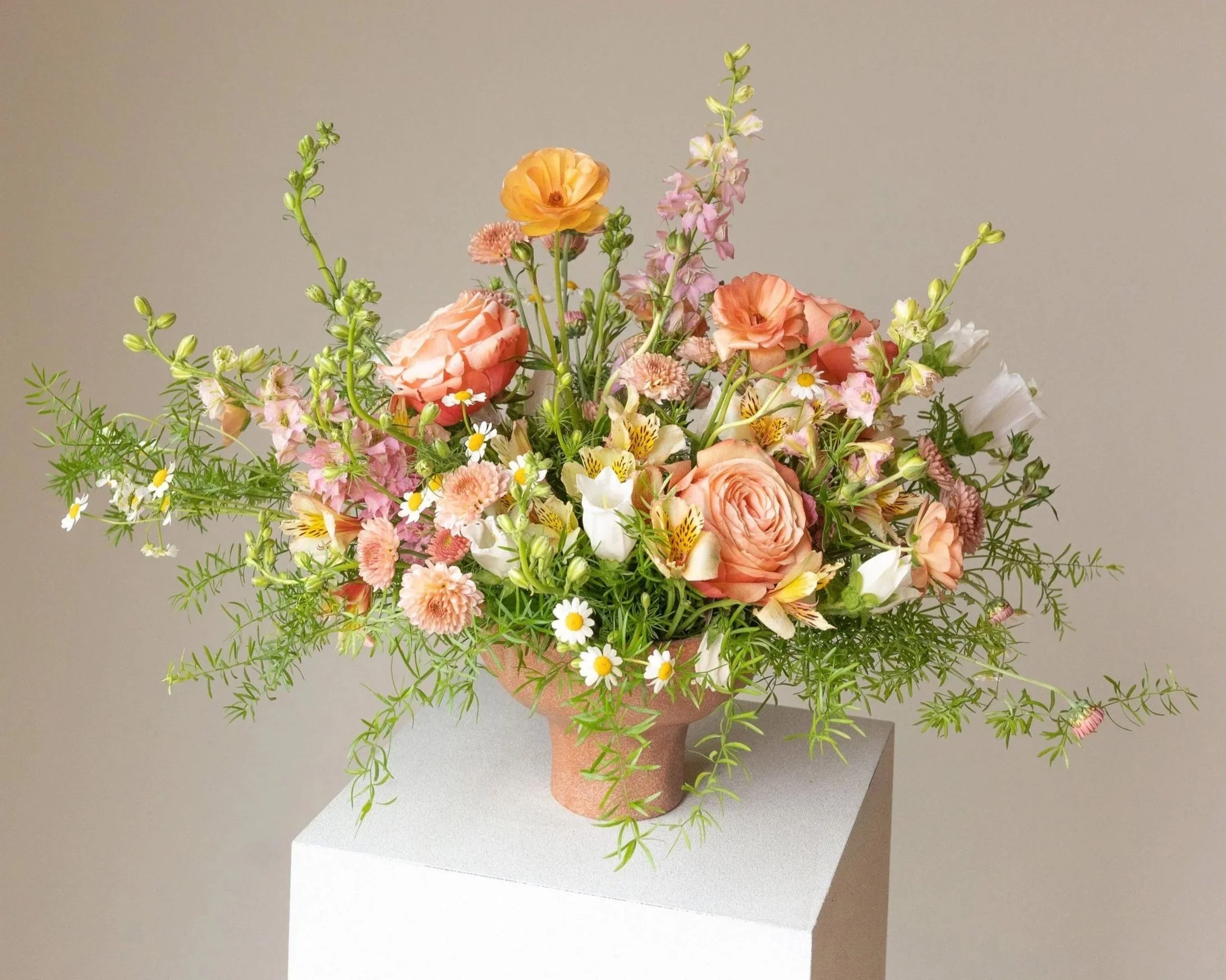 A colorful bouquet of various pink, white, and yellow flowers in a terracotta vase, placed on a white pedestal against a neutral background.