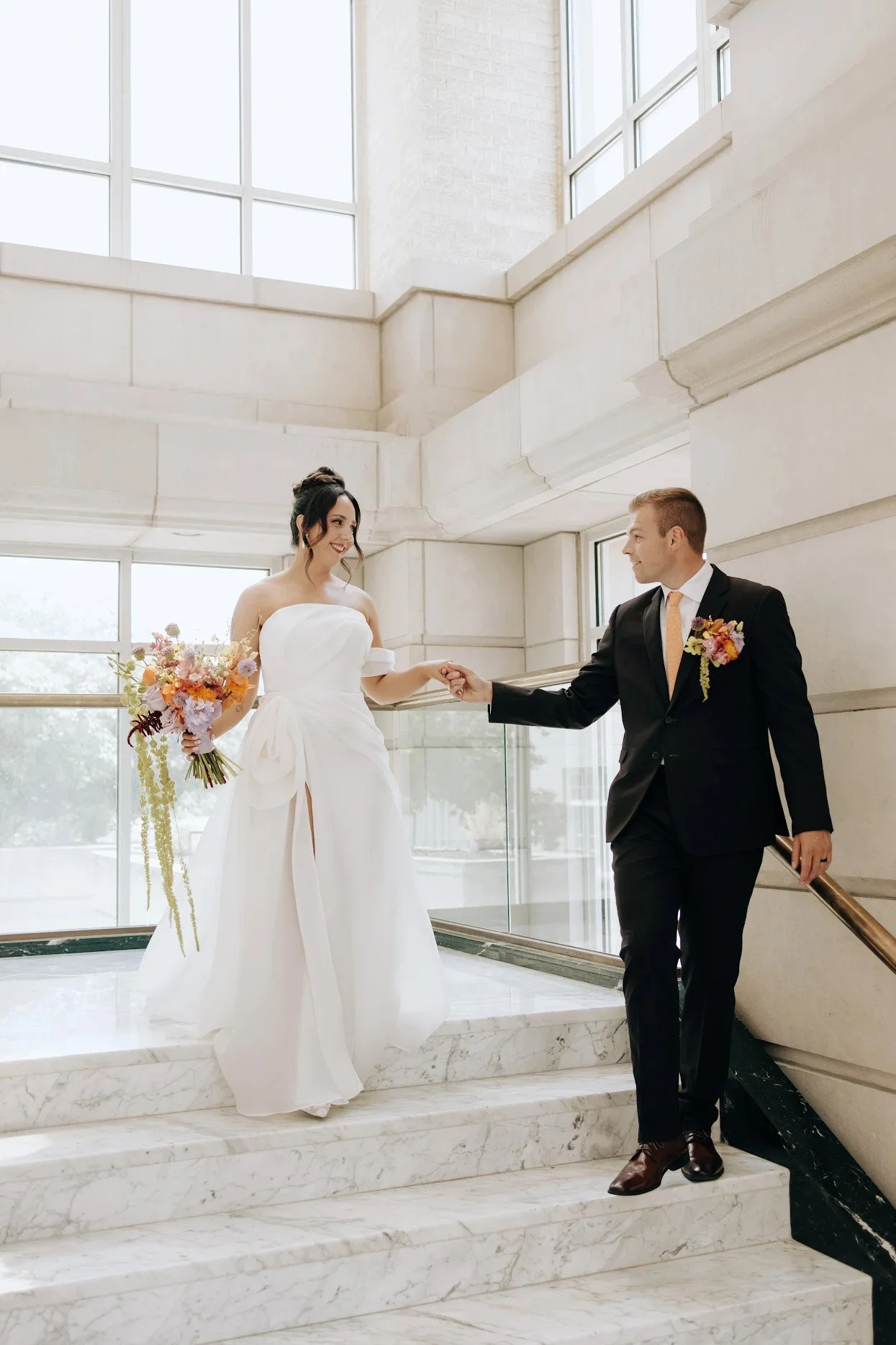 A bride in a white wedding gown holding a bouquet, holding hands with a groom in a black suit inside a modern building with large windows and marble stairs.