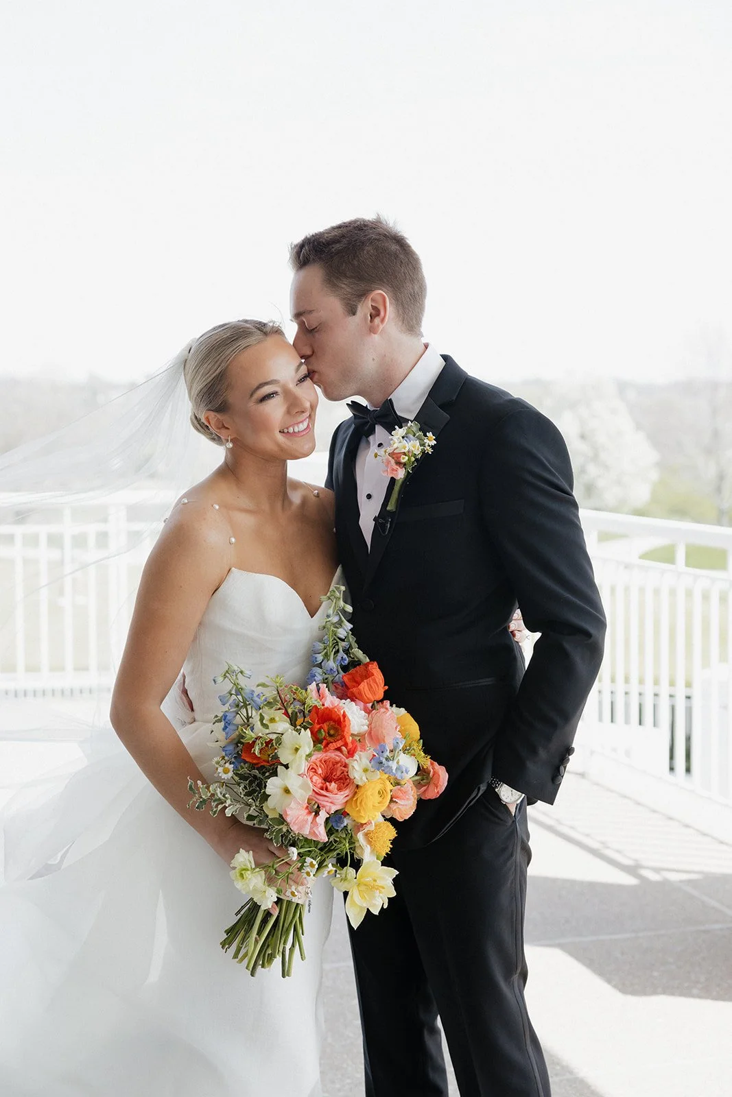 A bride and groom on their wedding day, with the groom kissing the bride on her forehead, both smiling, outdoors on a balcony, with the bride holding a colorful bouquet of flowers.