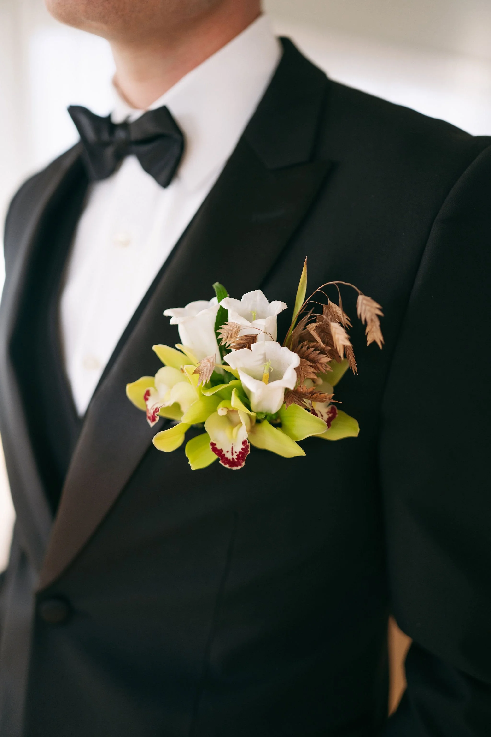 A man wearing a black tuxedo, white dress shirt, and black bow tie, with a boutonniere of white flowers and green leaves attached to his lapel.
