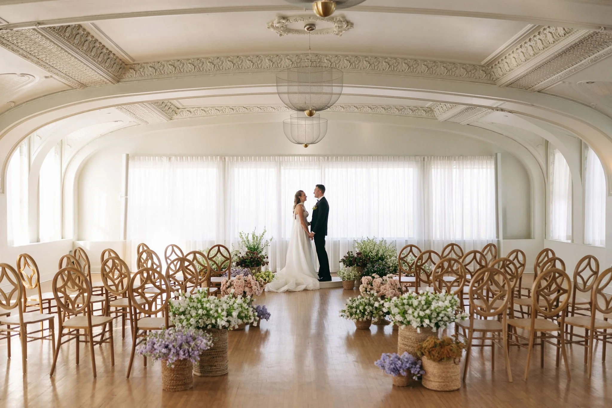 A bride and groom facing each other holding hands, standing on a small platform in a bright, ornate room with large windows, surrounded by floral arrangements and chairs arranged for a wedding ceremony.