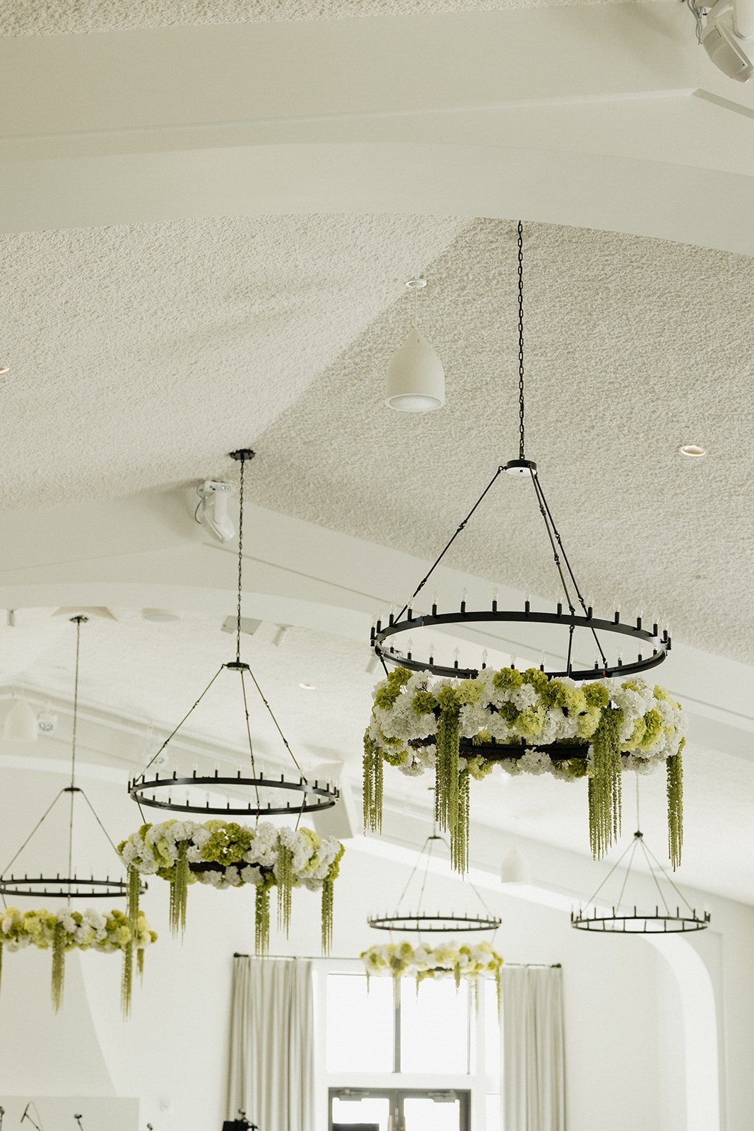 Chandeliers decorated with white and green flowers hanging from a white ceiling in a bright room.