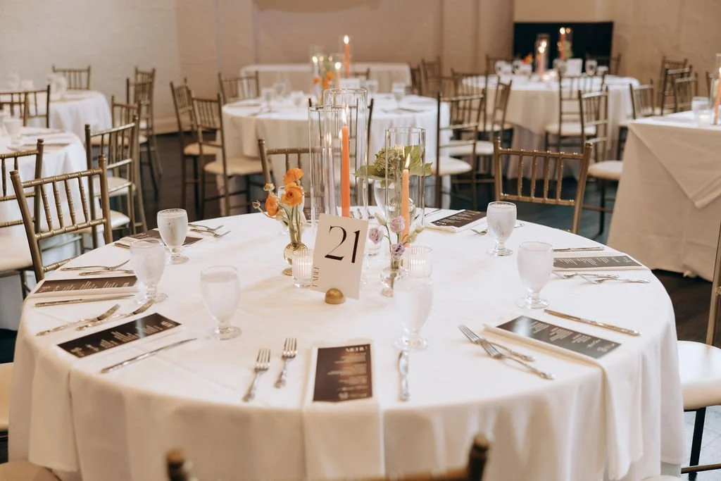 A banquet hall decorated for an event with round tables covered in white tablecloths, each set with glassware, silverware, and printed menus. The main table features tall candle holders with candles, small flower arrangements, and a table number card