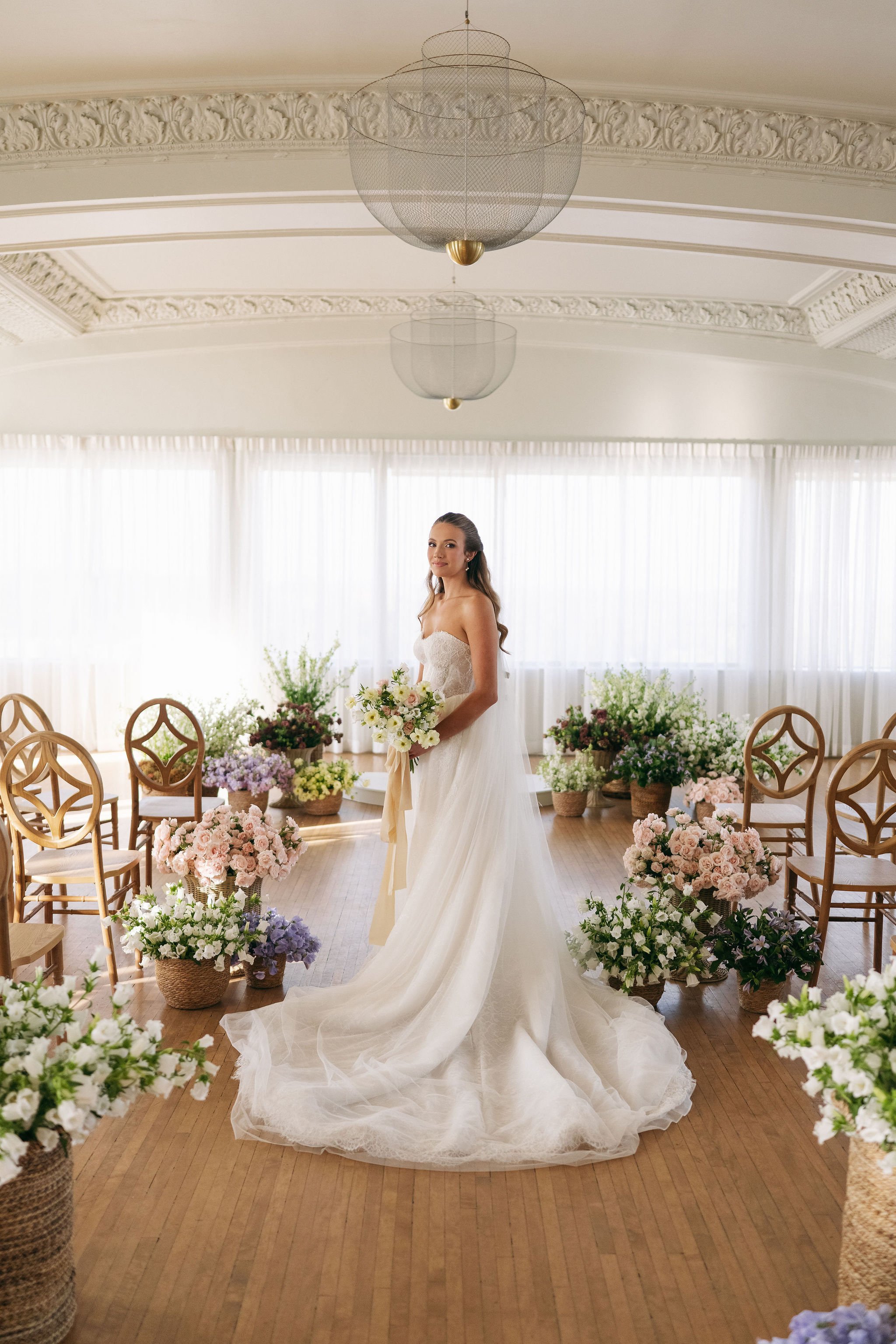A bride in a white wedding dress holding a bouquet standing in a decorated room filled with flowers and wooden chairs.