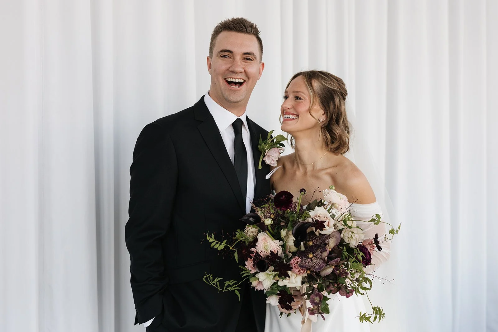 A newlywed couple smiling, with the groom in a black suit and tie, and the bride in a white off-shoulder dress holding a large bouquet of dark purple, blush pink, and white flowers, standing in front of a light-colored curtain.