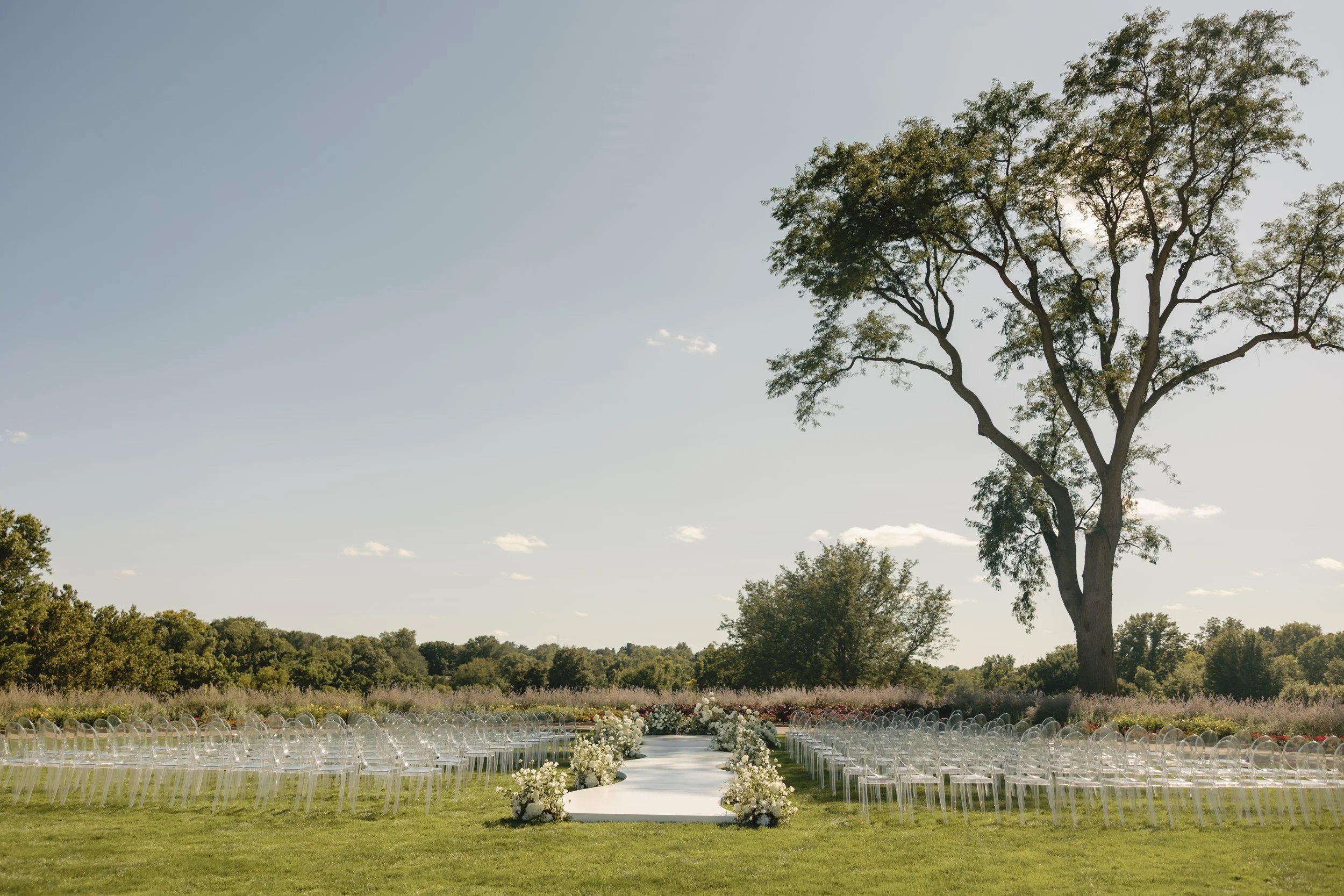 Outdoor wedding setup with a white aisle runner, surrounded by white floral arrangements, on a grassy field with a large tree and green trees in the background under a clear sky.